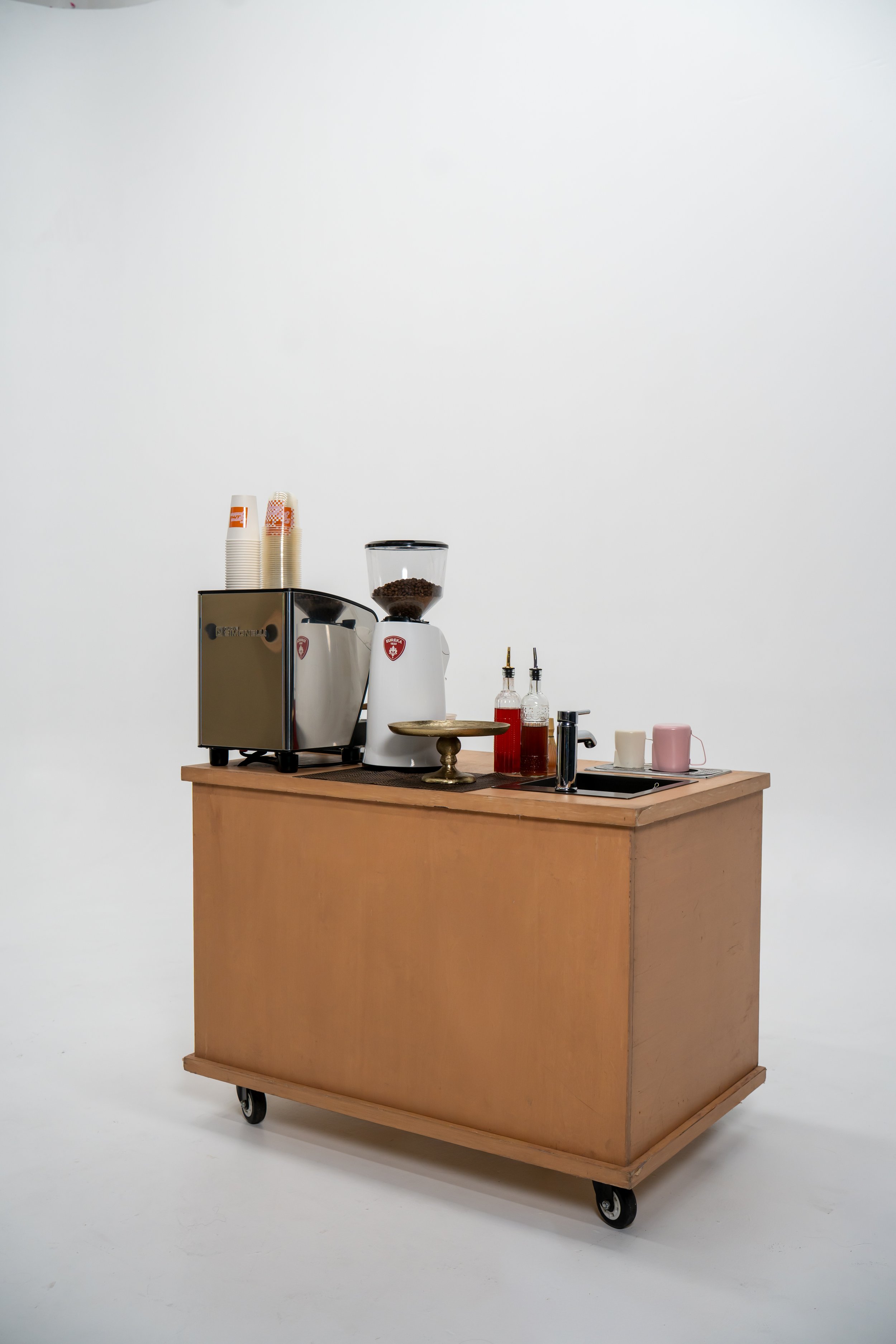 A coffee station on a wooden mobile cart with black wheels, set against a plain white wall. The setup includes an espresso machine, cups, and condiment bottles.