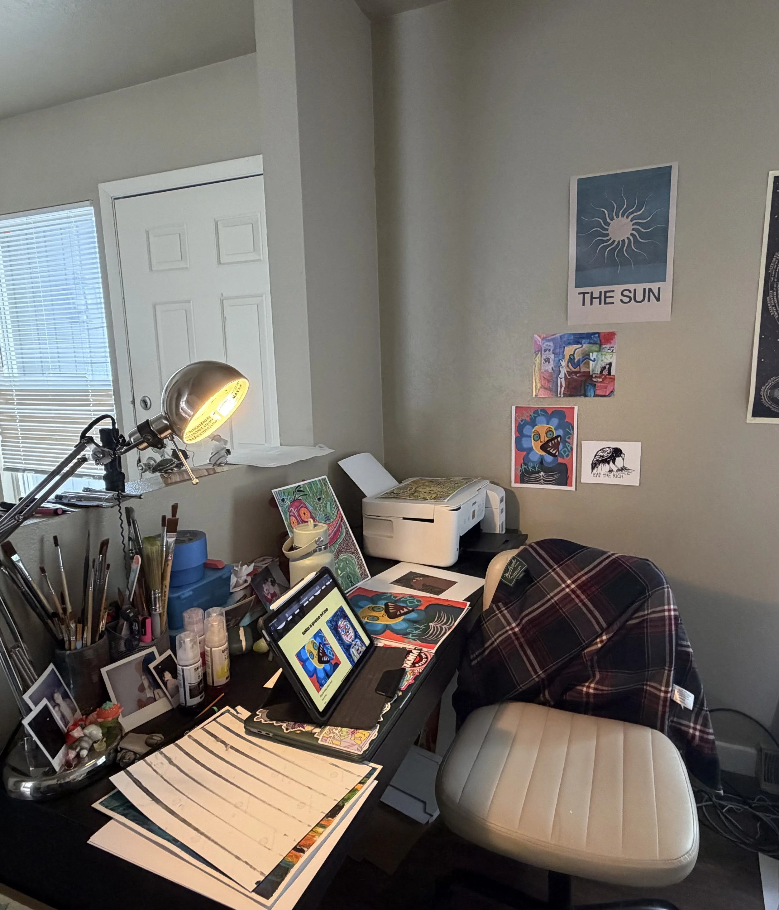 A desk in a room with various art supplies, printed artwork, and a tablet displaying colorful art. The wall has posters, including one labeled 'THE SUN,' and a photo of colorful abstract art. There's a plaid chair and a window with blinds.