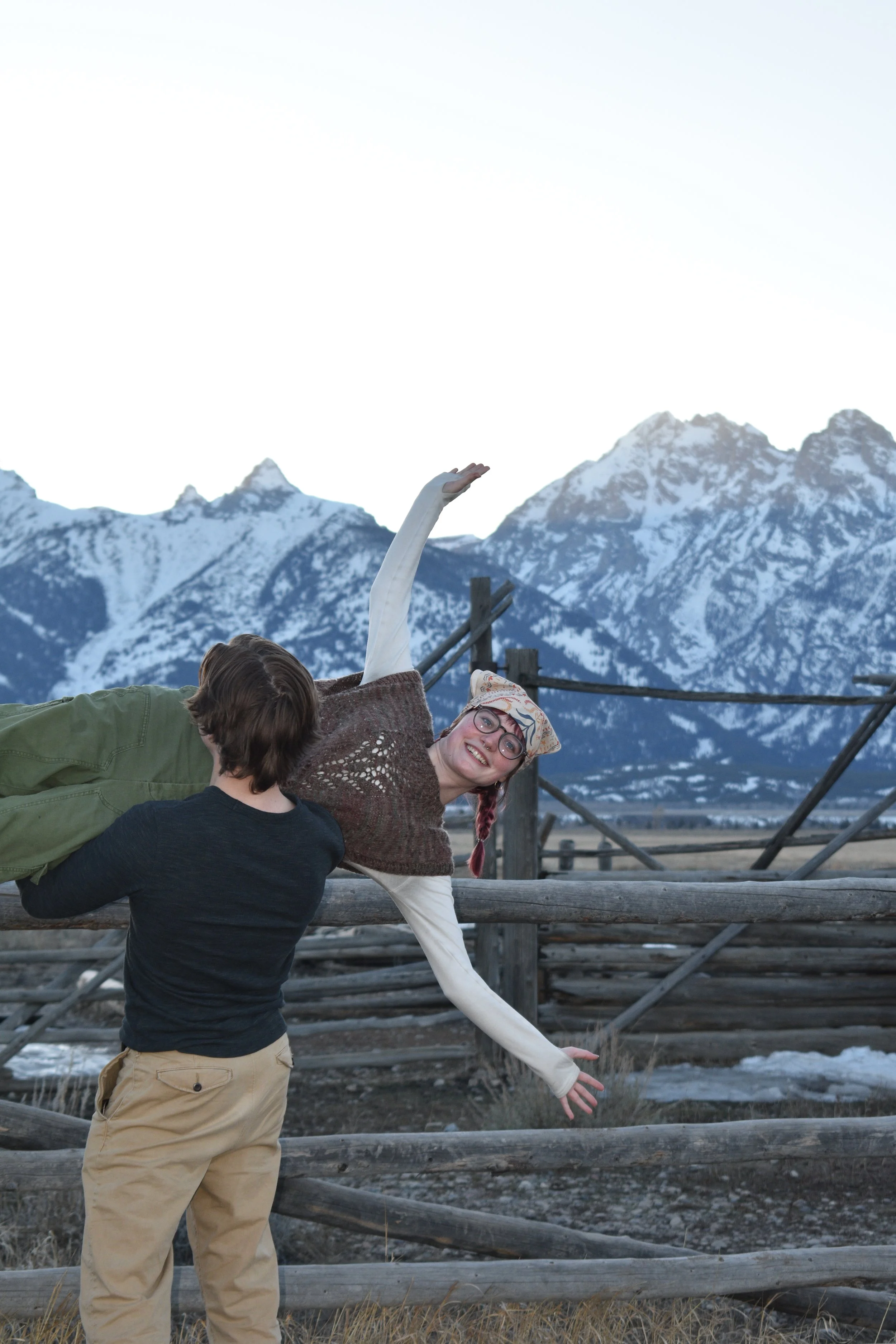 Three people outdoors near a mountain range with snow, standing on a wooden fence, one person raising an arm, and two others facing away.