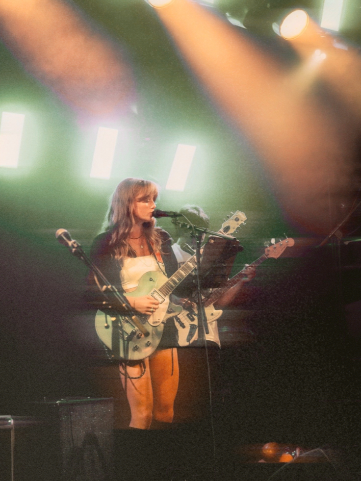 A woman with long hair is performing on stage, playing an electric guitar and singing into a microphone. Stage lights and equipment are visible in the background.