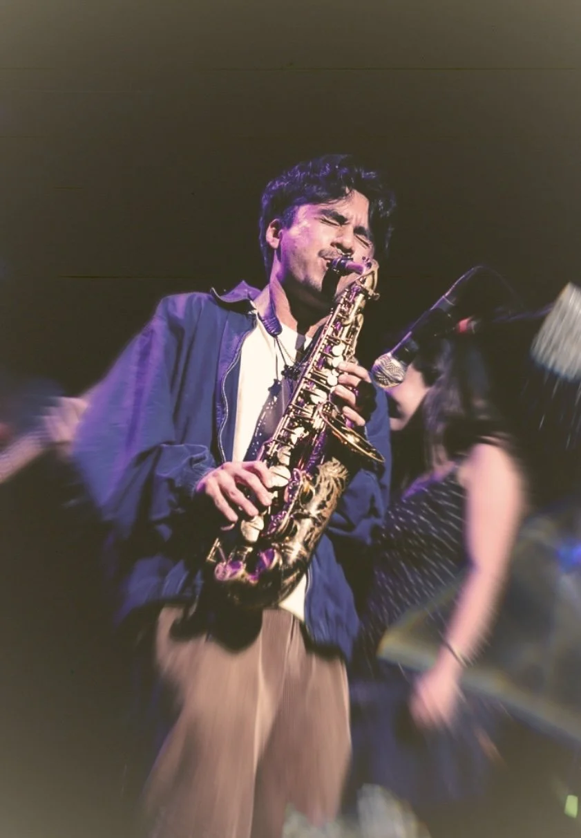 A man passionately playing an alto saxophone on stage, eyes closed, with people in the background.