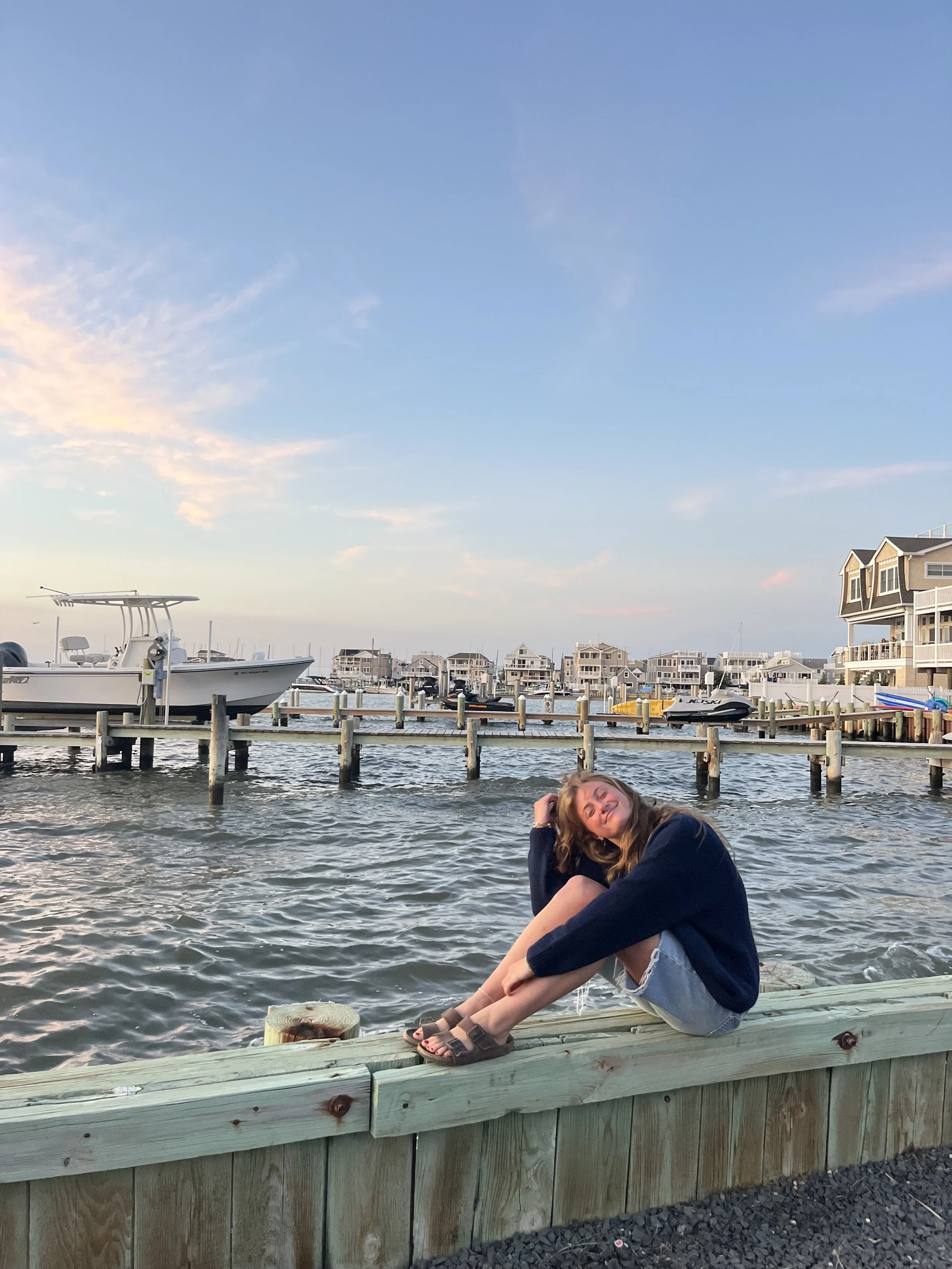 A young woman sitting on a wooden dock near the water, with boats docked in the background and a sky with scattered clouds.