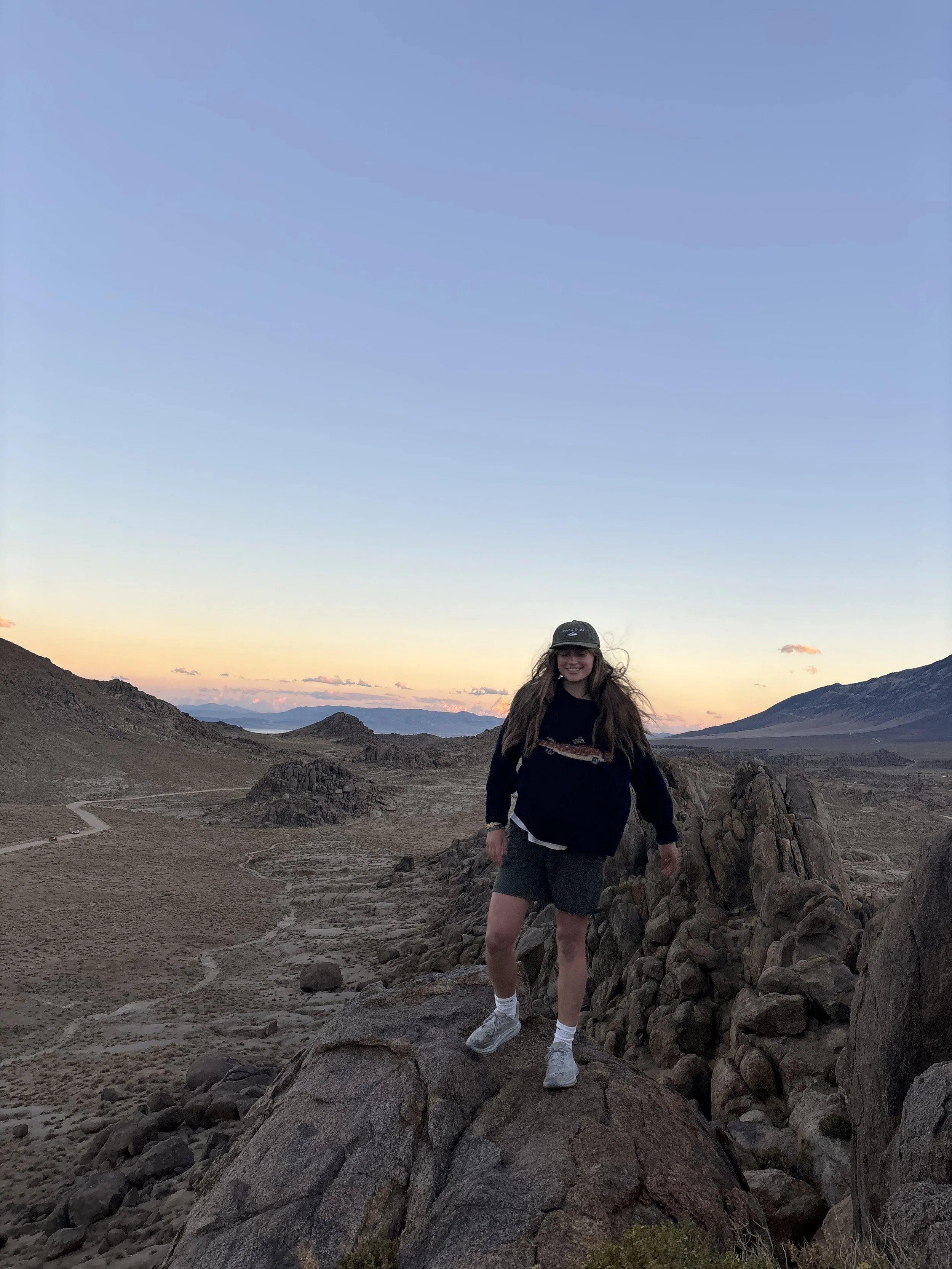 A woman with long hair wearing a hat, dark sweatshirt, shorts, and sneakers, standing on rocks in a desert landscape with mountains in the background during sunset.