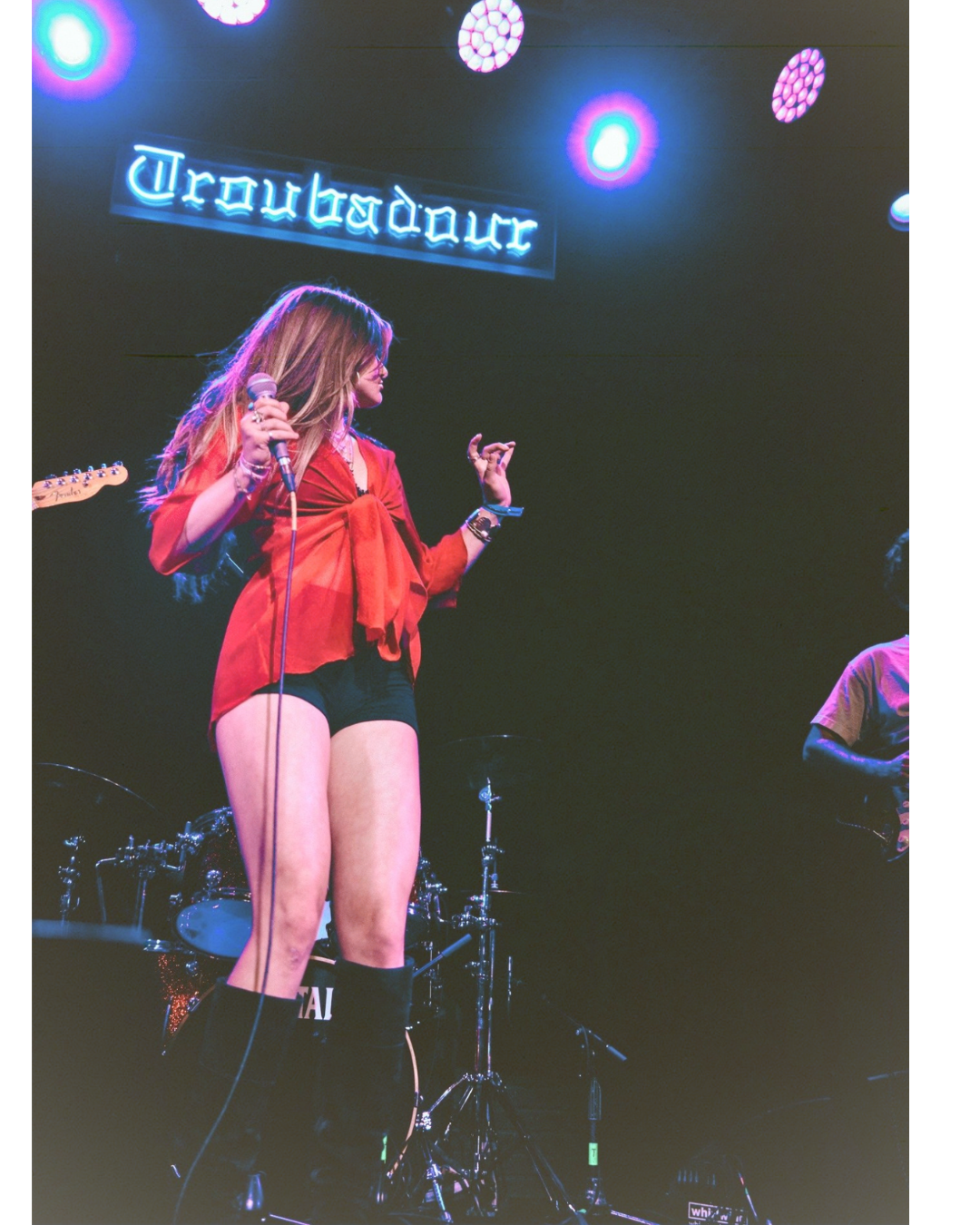 A woman performing on stage at Troubadour venue, wearing a red jacket, black shorts, and knee-high socks, holding a microphone in her right hand.