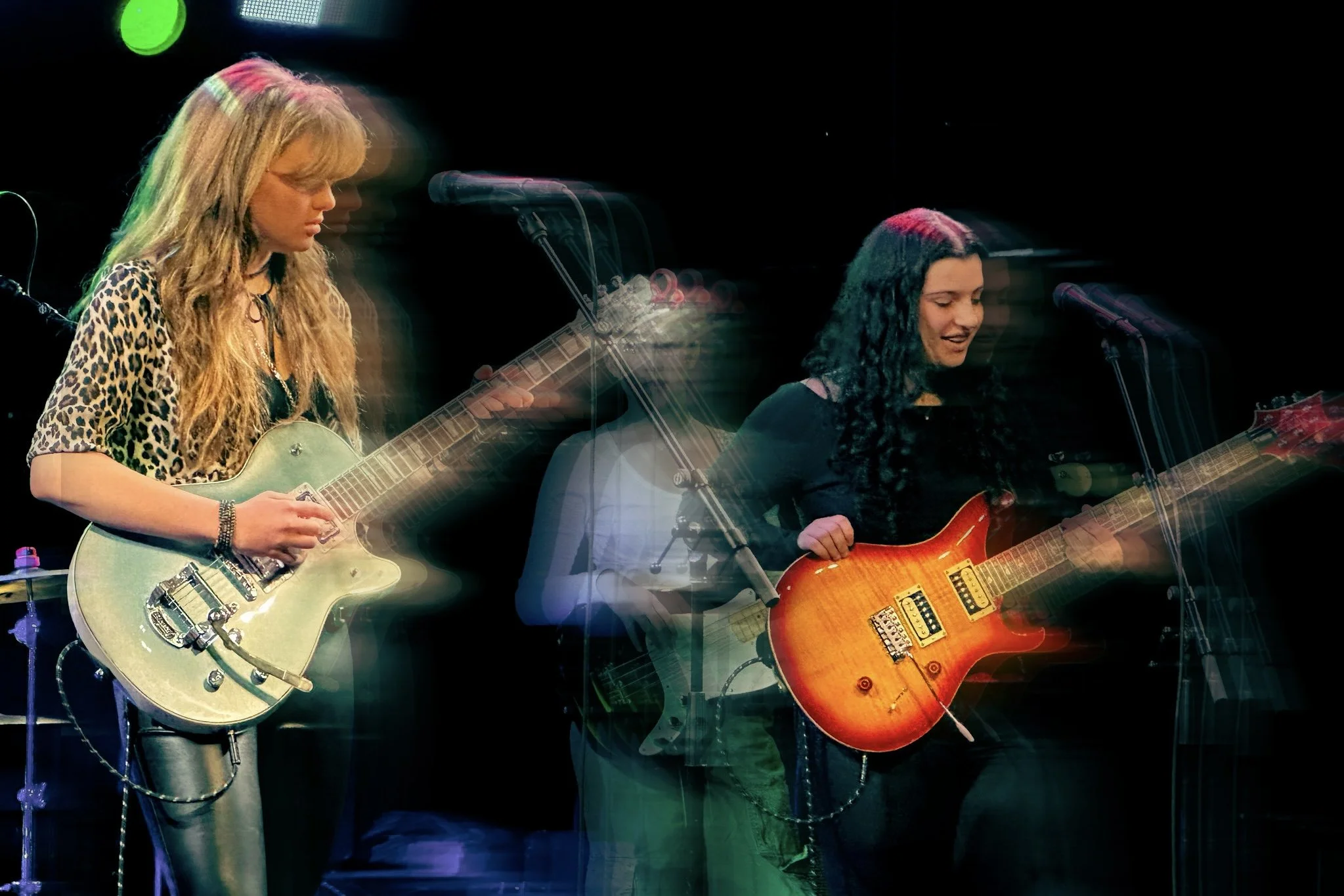 Two female musicians performing on stage, playing guitars with a black background and stage lighting.