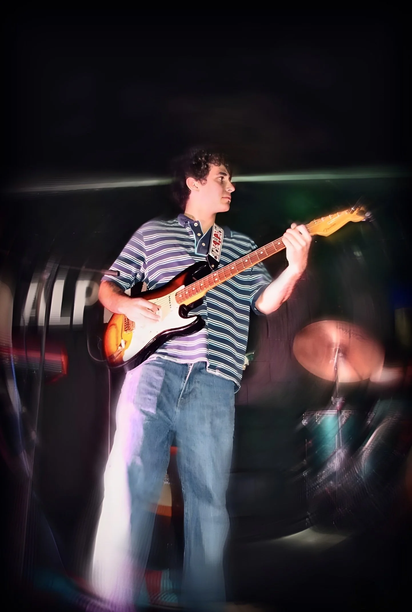 A young man with curly hair wearing a striped polo shirt and jeans playing an electric guitar on a stage with a dark background and colorful stage lights.