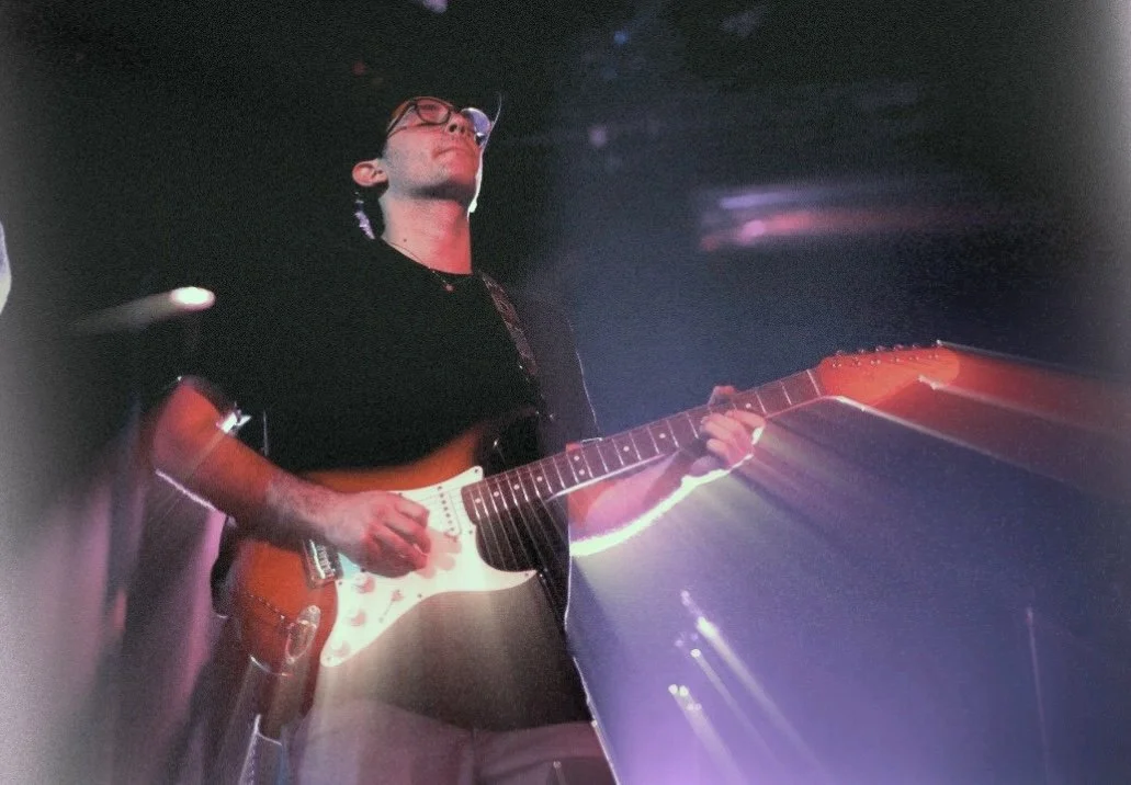 A person wearing glasses is playing an electric guitar on a dark stage with colorful lights.