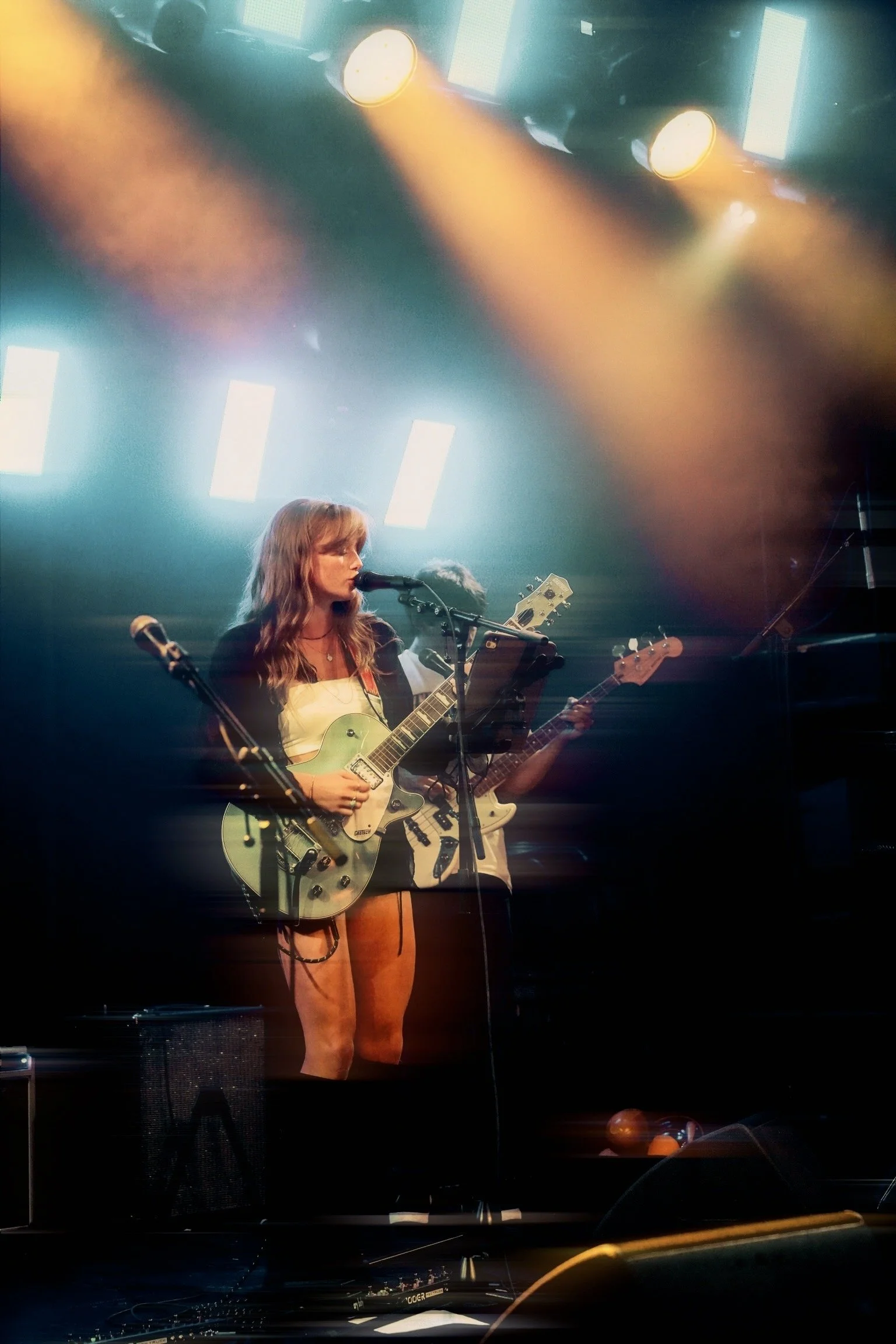 A woman performing on stage, playing a guitar and singing into a microphone, with colorful stage lights overhead.