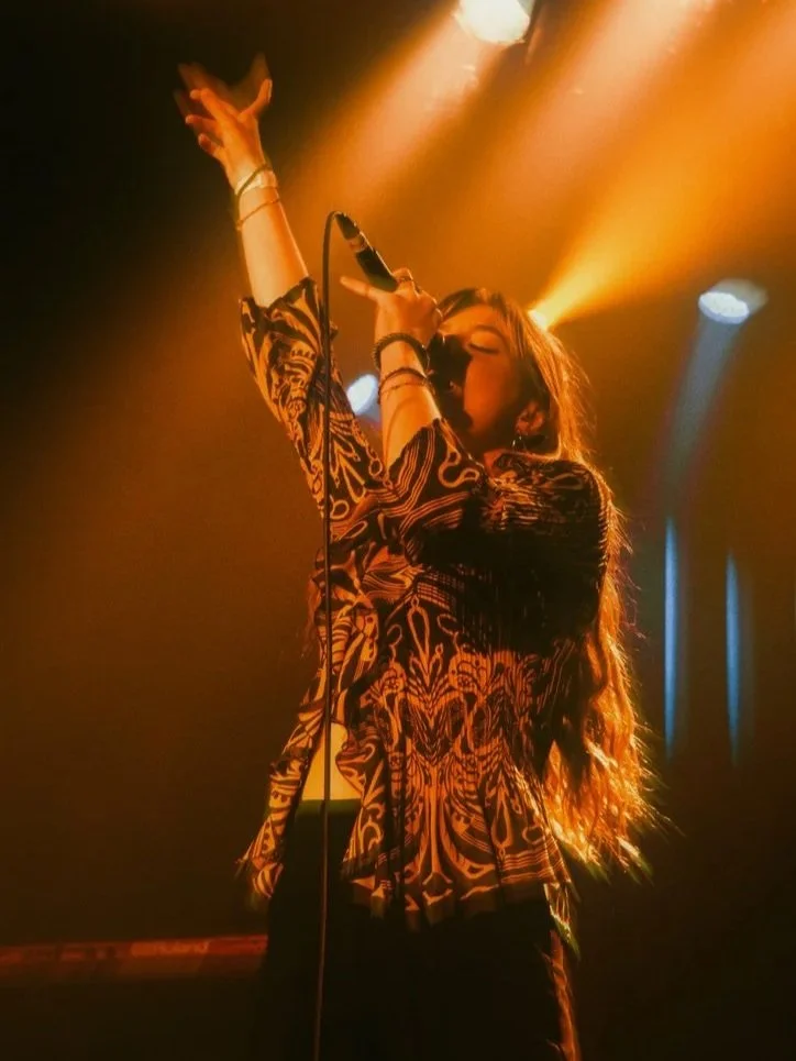 A female singer performing on stage, holding a microphone with one hand raised, under dramatic orange and blue stage lights.