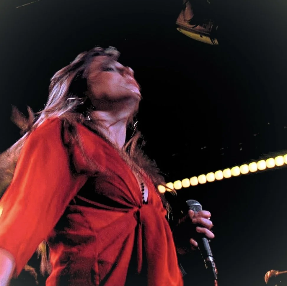 A woman singing into a microphone on stage, wearing a red jacket, with stage lights in the background.