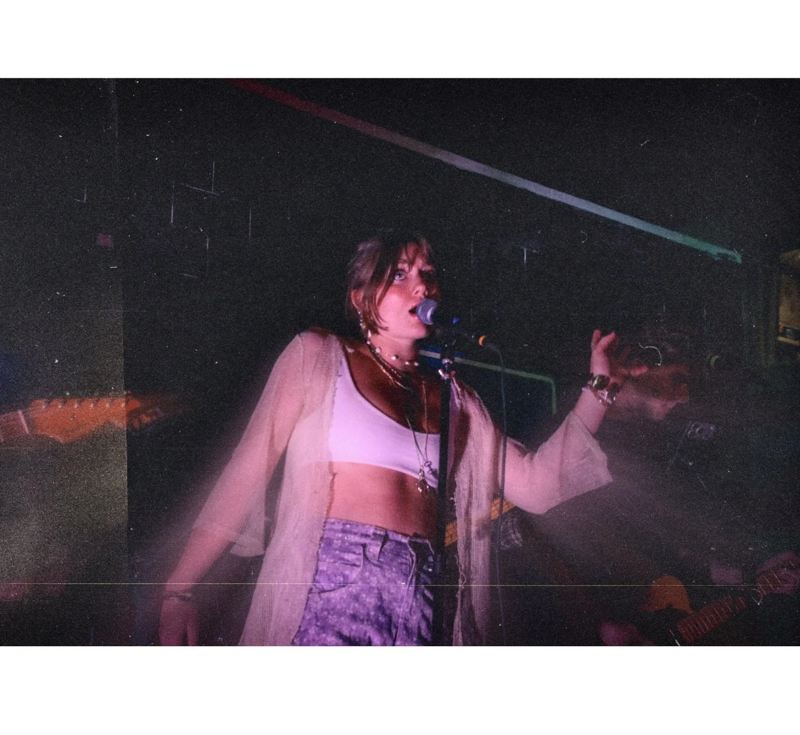 A woman singing into a microphone on stage with a colorful light spectrum above her. She is wearing a crop top, skirt, and jacket, with jewelry and accessories. A person playing guitar is visible in the background.