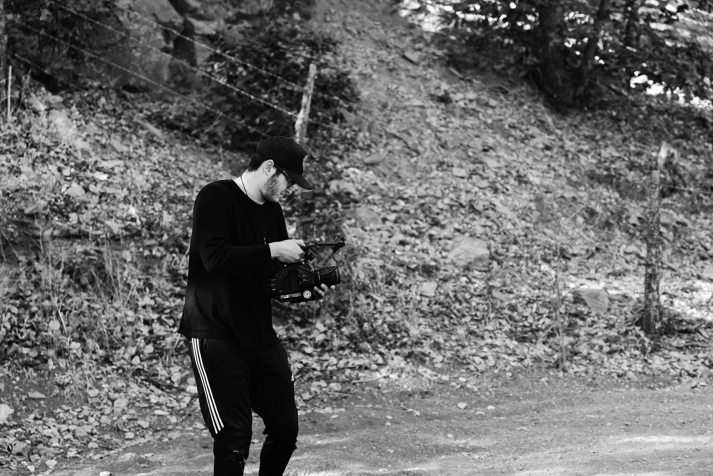 A young man wearing a black cap, glasses, and a black long-sleeve shirt stands outdoors in a wooded area holding a camera on a stabilizer. The background features a hillside with trees, rocks, and a barbed wire fence.