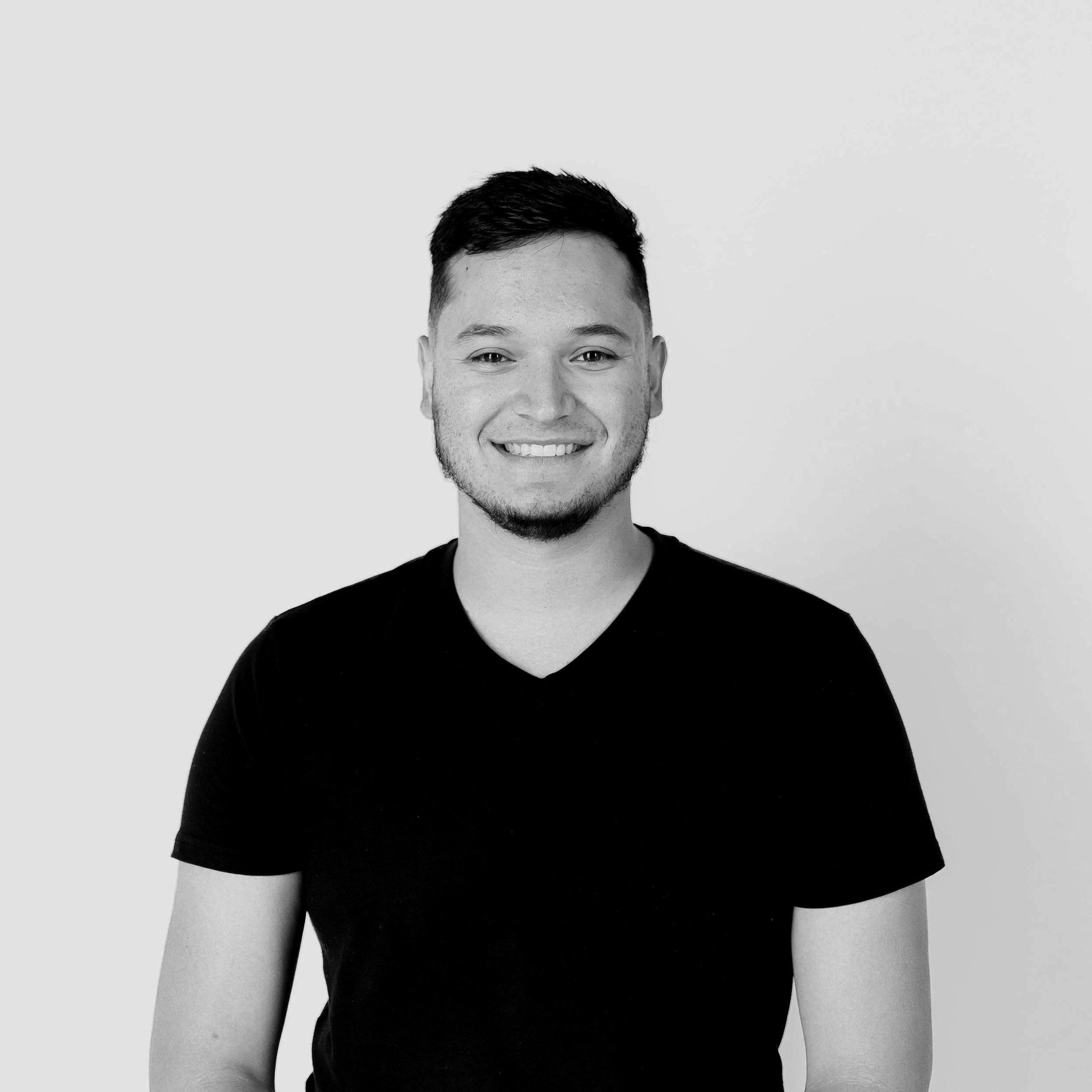 Black and white photo of a young man smiling, wearing a dark V-neck T-shirt, against a plain white background. (He's the Founder and CEO)