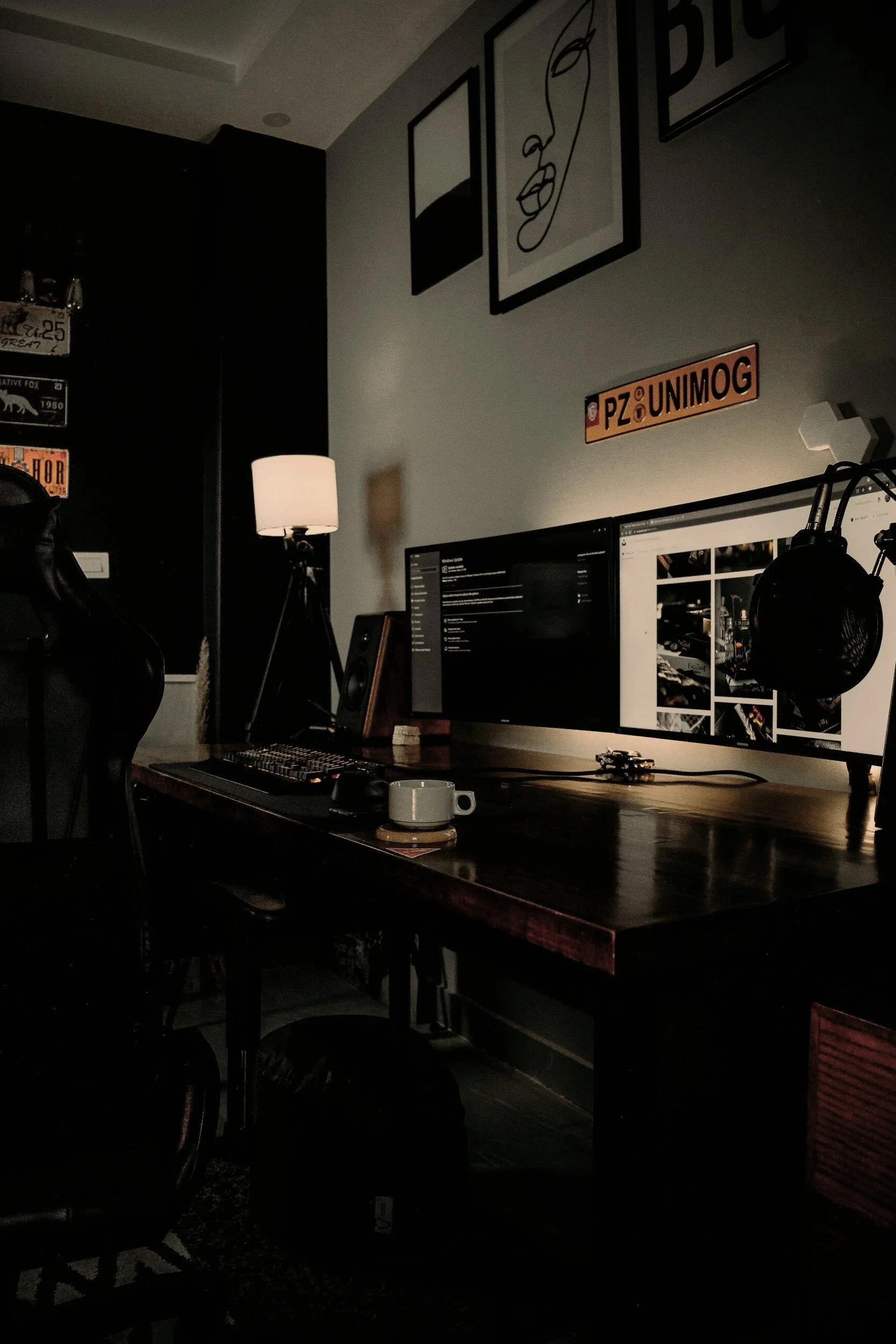 A computer workspace in a dimly lit room with a wooden desk, dual monitors displaying a website, a keyboard, a mouse, a coffee cup, and a microphone. The wall has framed artworks, vintage license plates, and signage.