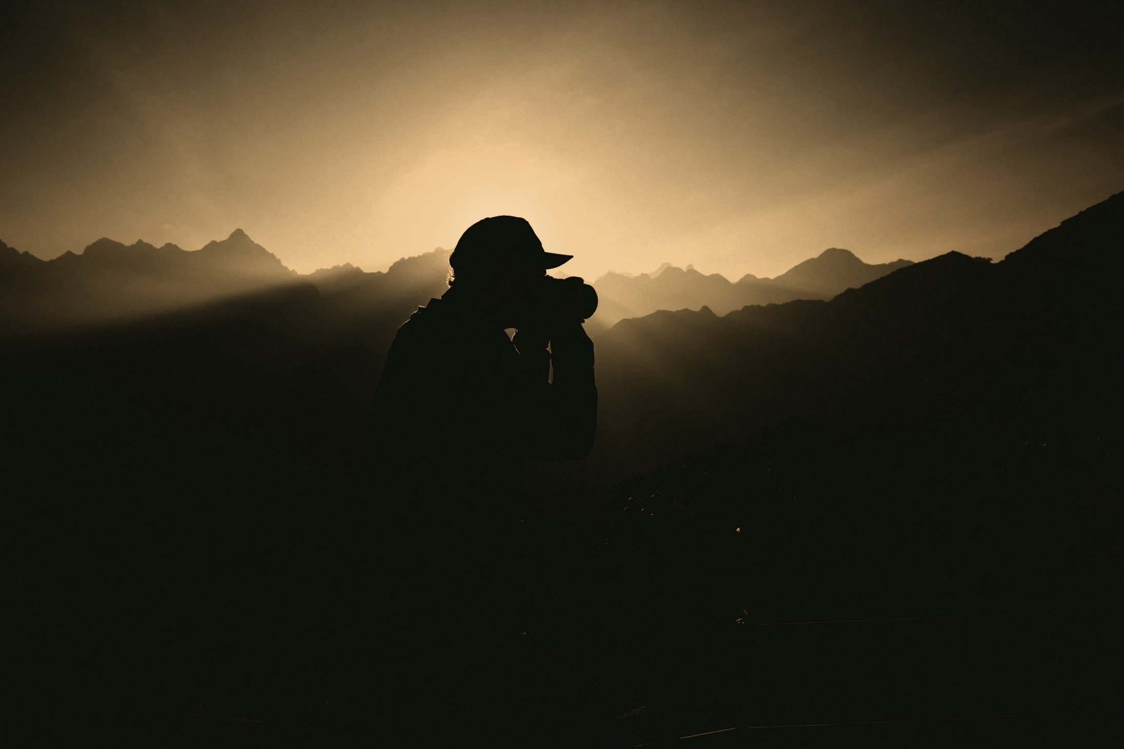 Silhouette of a person with a hat, holding a camera, standing in front of mountains at sunset or sunrise (which symbolizes creative sollutions).