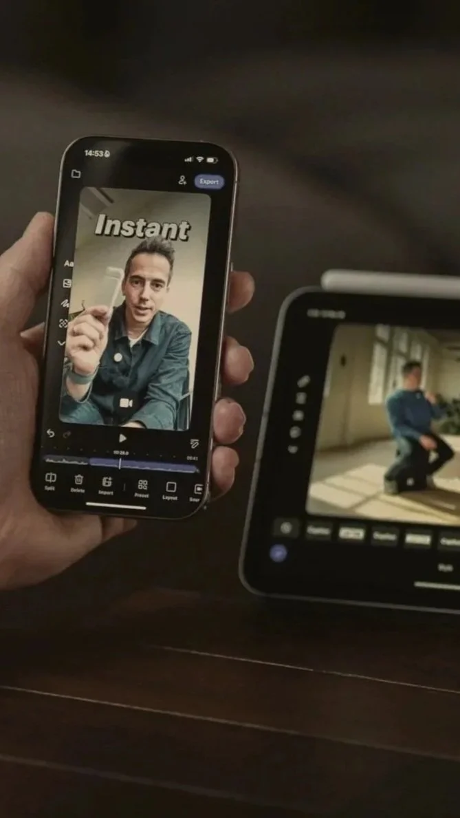 Person holding a smartphone showing a video editing app with a man gesturing and the word "Instant" on the screen. There is a tablet on the table displaying a video of a person kneeling on the floor.