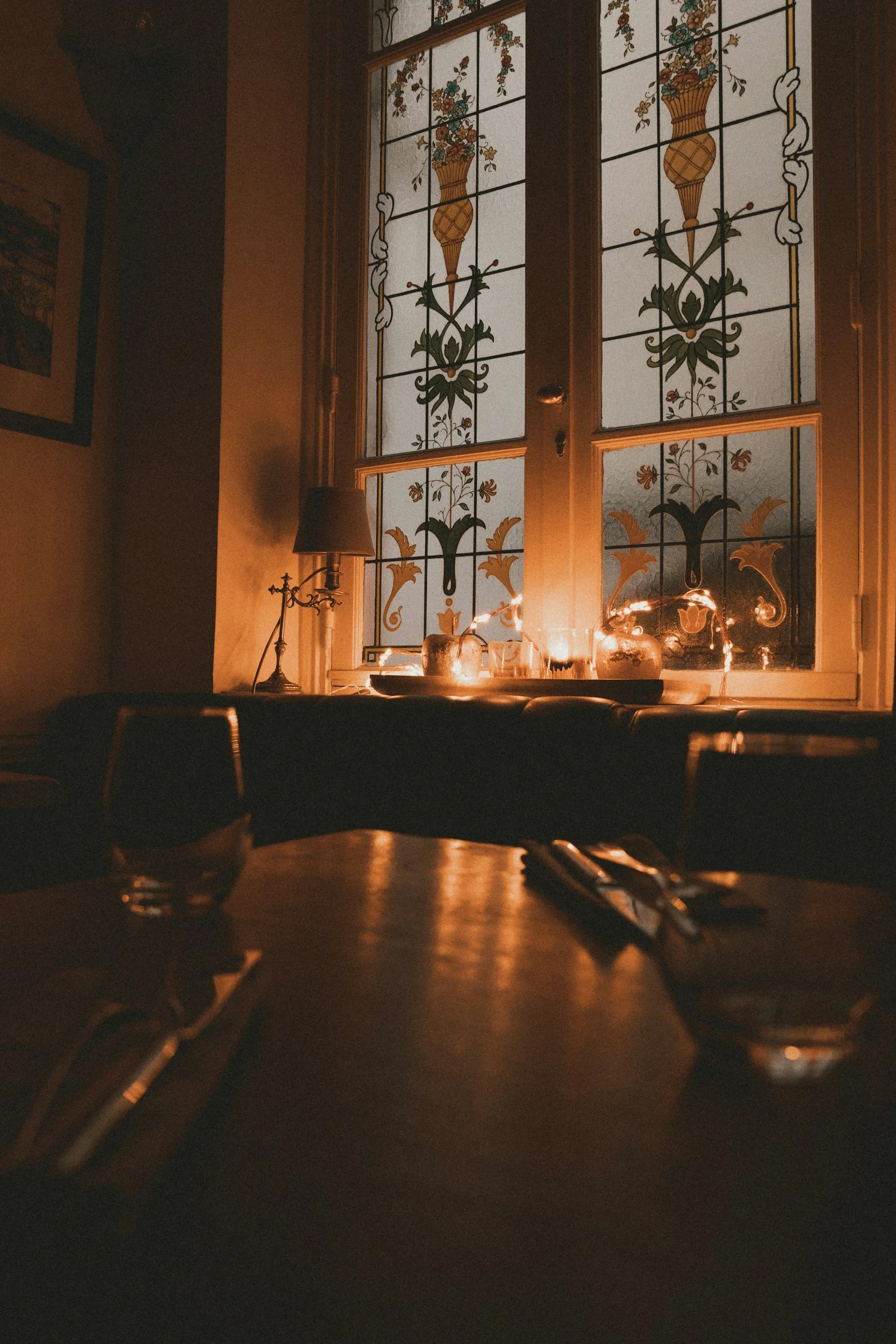 A cozy indoor scene featuring stained glass window panels with floral and geometric patterns, a table with glasses and candles, a small lamp, and dim lighting creating a warm atmosphere for an intimate anniversary dinner celebration in Nashville.