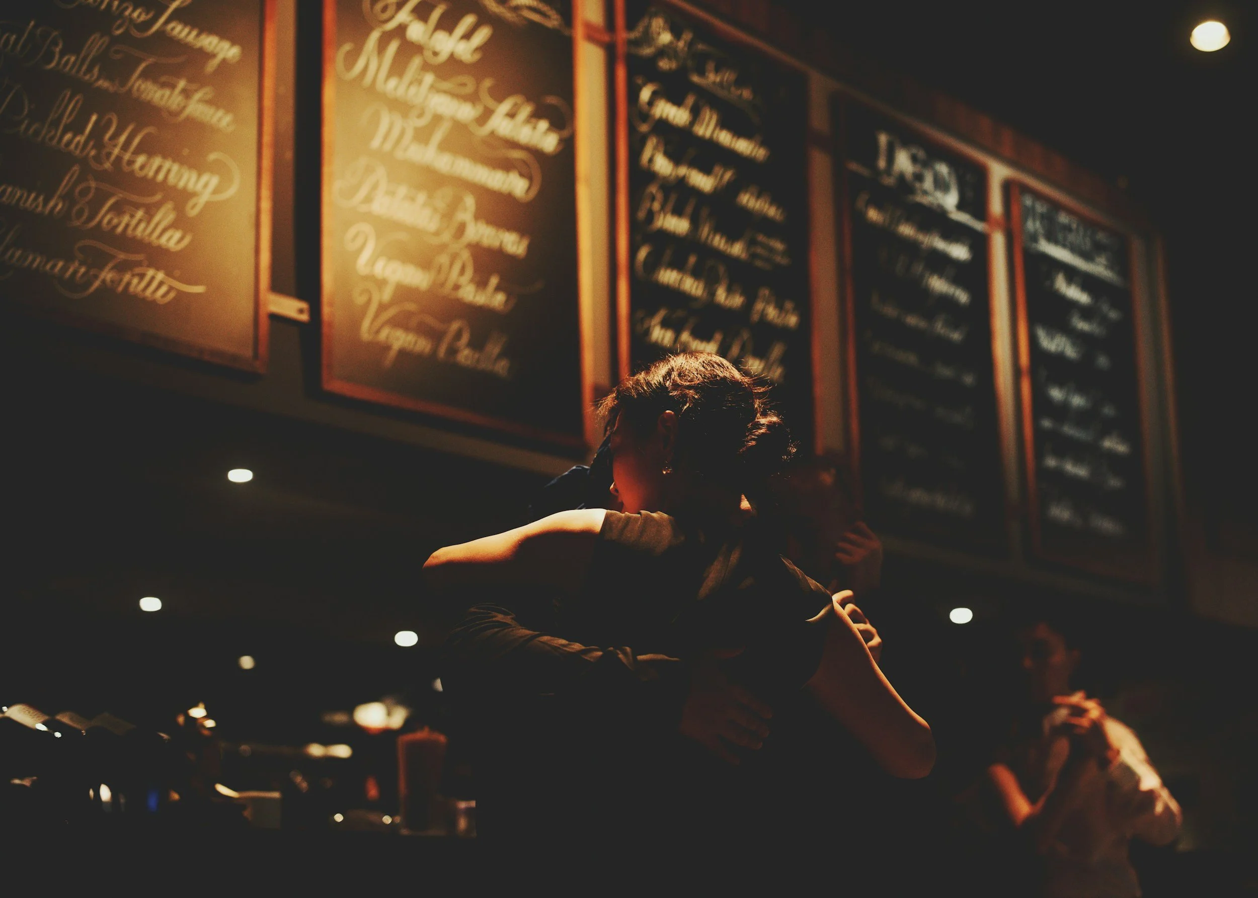 A couple hugging in a dimly lit indoor setting, with a blackboard menu with gold lettering in the background.