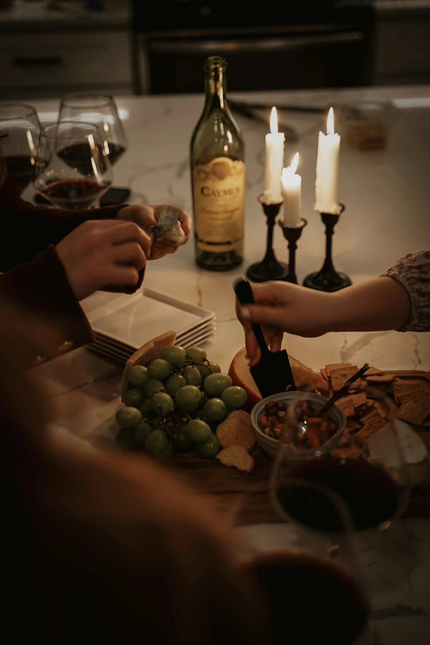 A dinner table with three lit white candles, a wine bottle, several glasses of red wine, a bunch of green grapes, cheese, crackers, and a bowl of mixed nuts or snacks.
