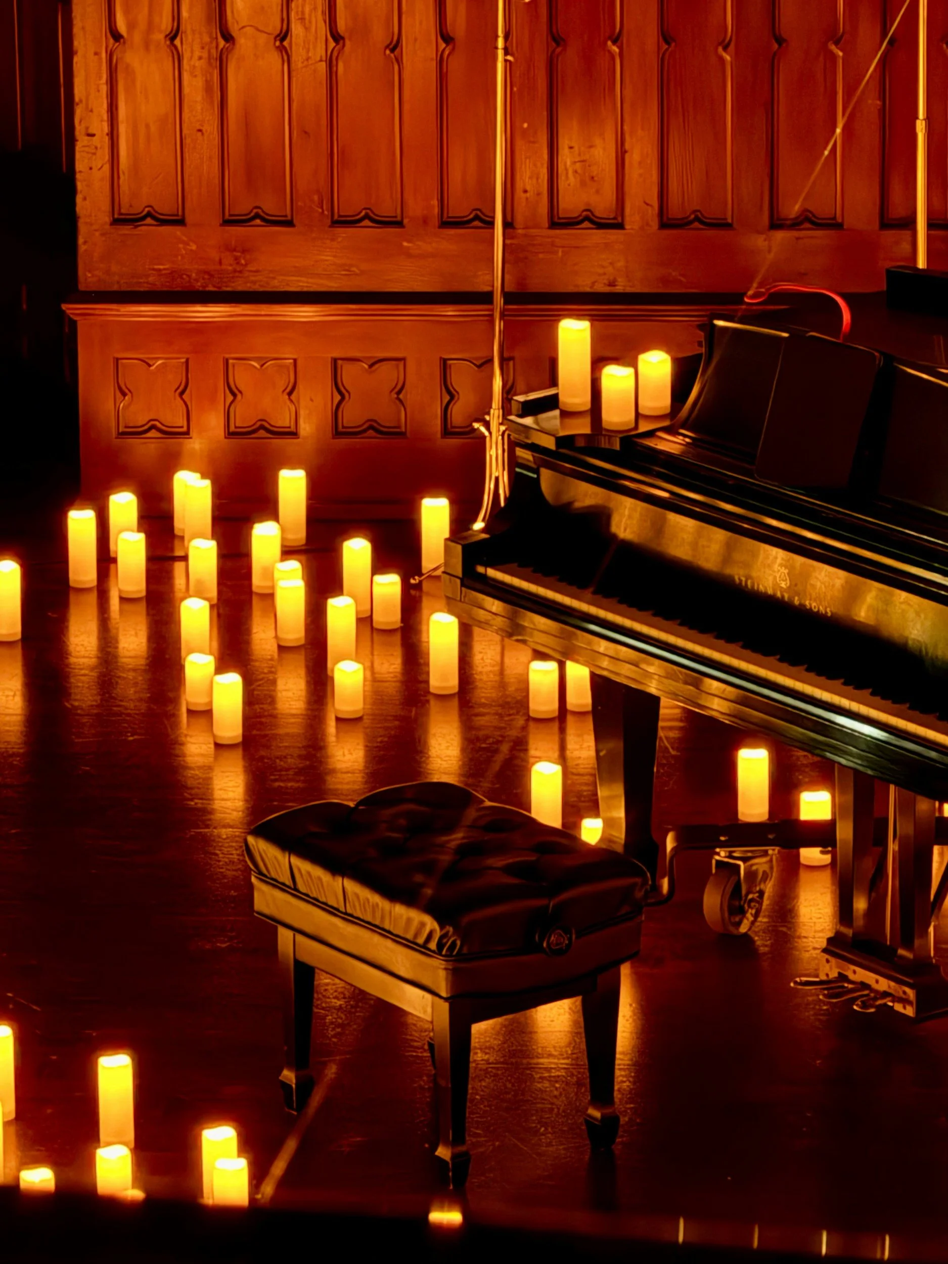 An intimate anniversary celebration featuring a grand piano with a glossy black finish sits in a dimly lit room illuminated by numerous yellow candles, some on the piano and some on the floor. The background features wooden paneling on the wall.
