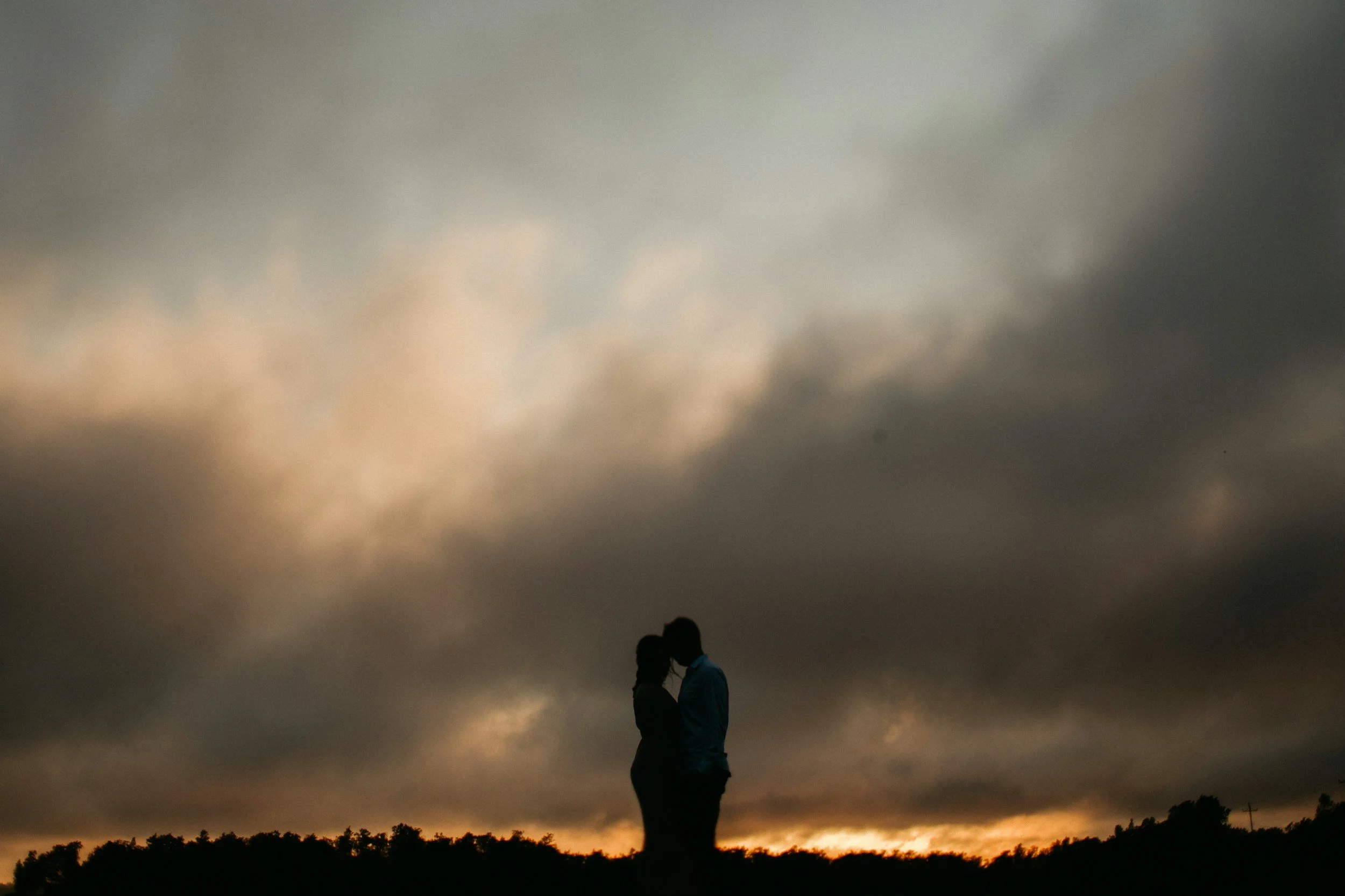 A silhouette of a couple standing in an embrace while outdoors at sunset or dusk, with dark clouds overhead and a line of trees or shrubs on the horizon.