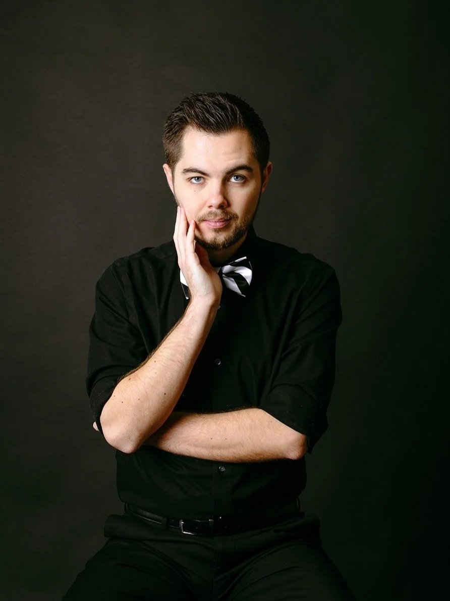 Mentalist Mark Gibson, wearing a black shirt and a black and white striped bow tie, posing against a dark background with one hand touching his cheek and the other arm crossed.