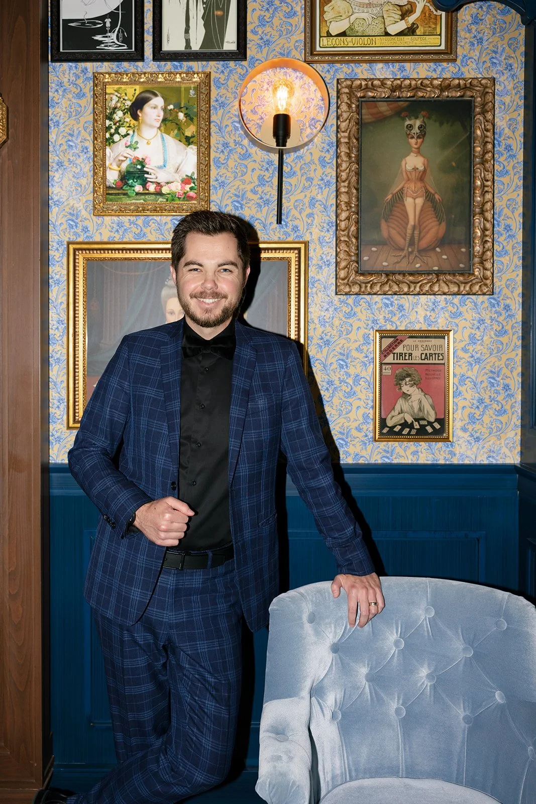Mentalist Mark Gibson in a blue checkered suit stands in a room with ornate wallpaper and various framed artworks on the wall, smiling at the camera.