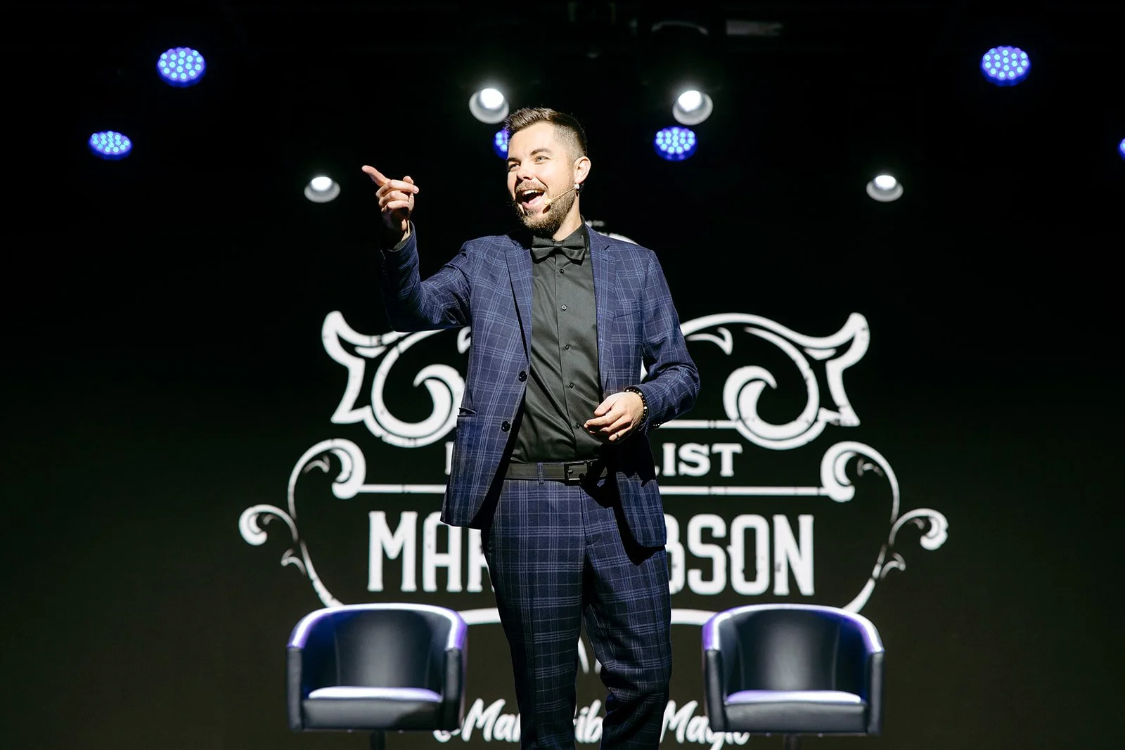 Mentalist Mark Gibson in a plaid suit and black shirt, with a microphone headset, is standing on a stage. He is smiling and pointing, with a stylized logo and the words 'MARK GIBSON MENTALIST' visible in the background.