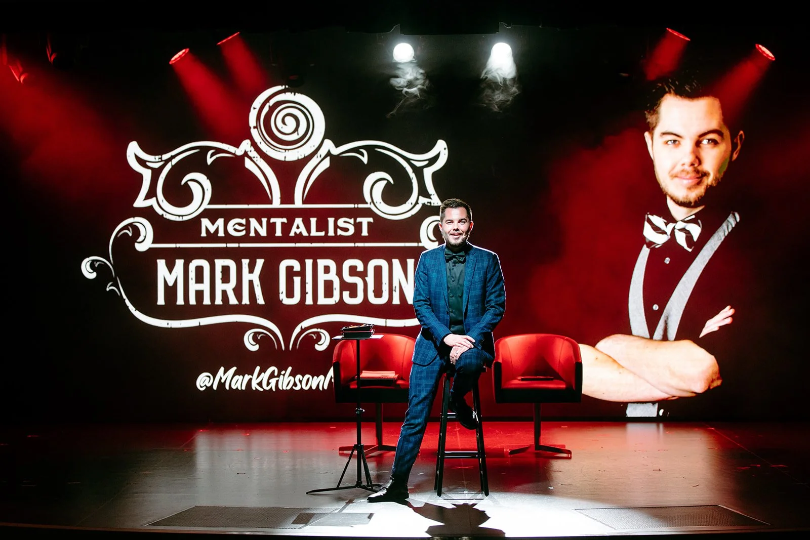 Mentalist Mark Gibson in a blue checkered suit sitting on a tall stool on stage, with two red chairs and a small table. Behind him is a large backdrop displaying the words 'MENTALIST MARK GIBSON' and instagram  '@MarkGibsonMagic. '