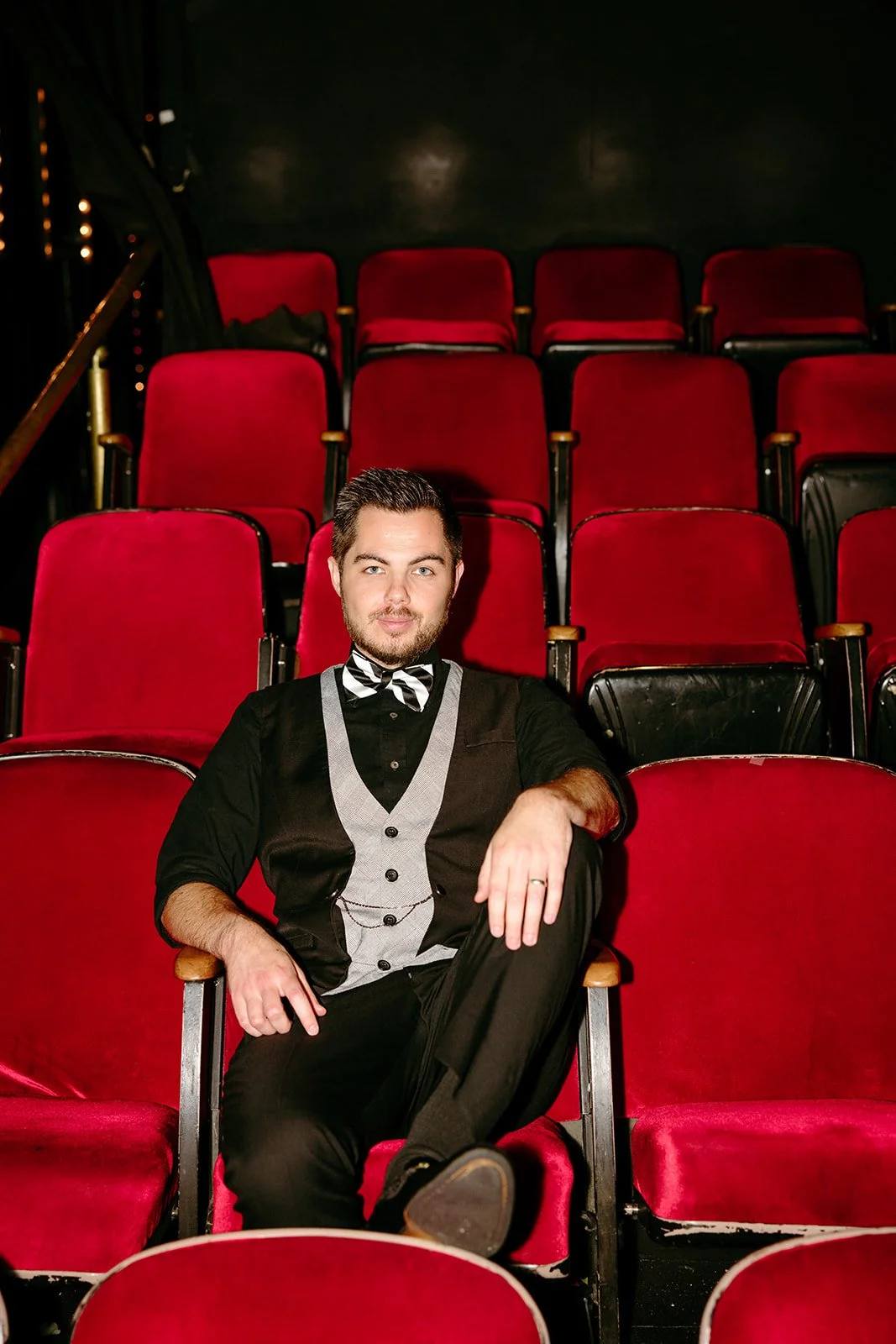 A man dressed in formal attire with a black shirt, gray vest, and striped bow tie sitting in an empty red theater seat.