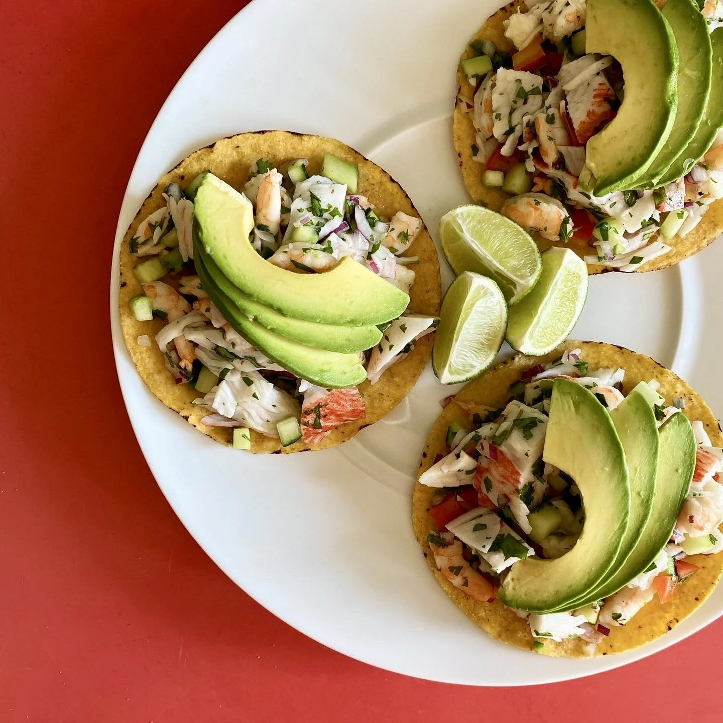 Three Mexican street corn tostadas topped with diced vegetables and sliced avocado, served with lime wedges on a white plate.