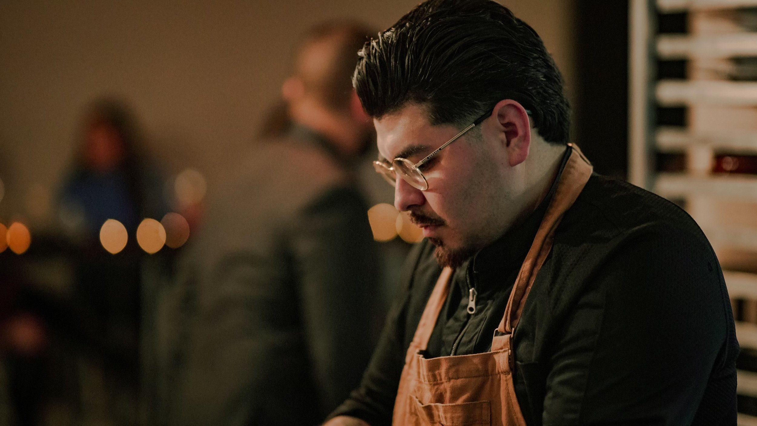 A man with dark hair, glasses, and a beard, wearing a brown apron, working in a dimly lit indoor setting.