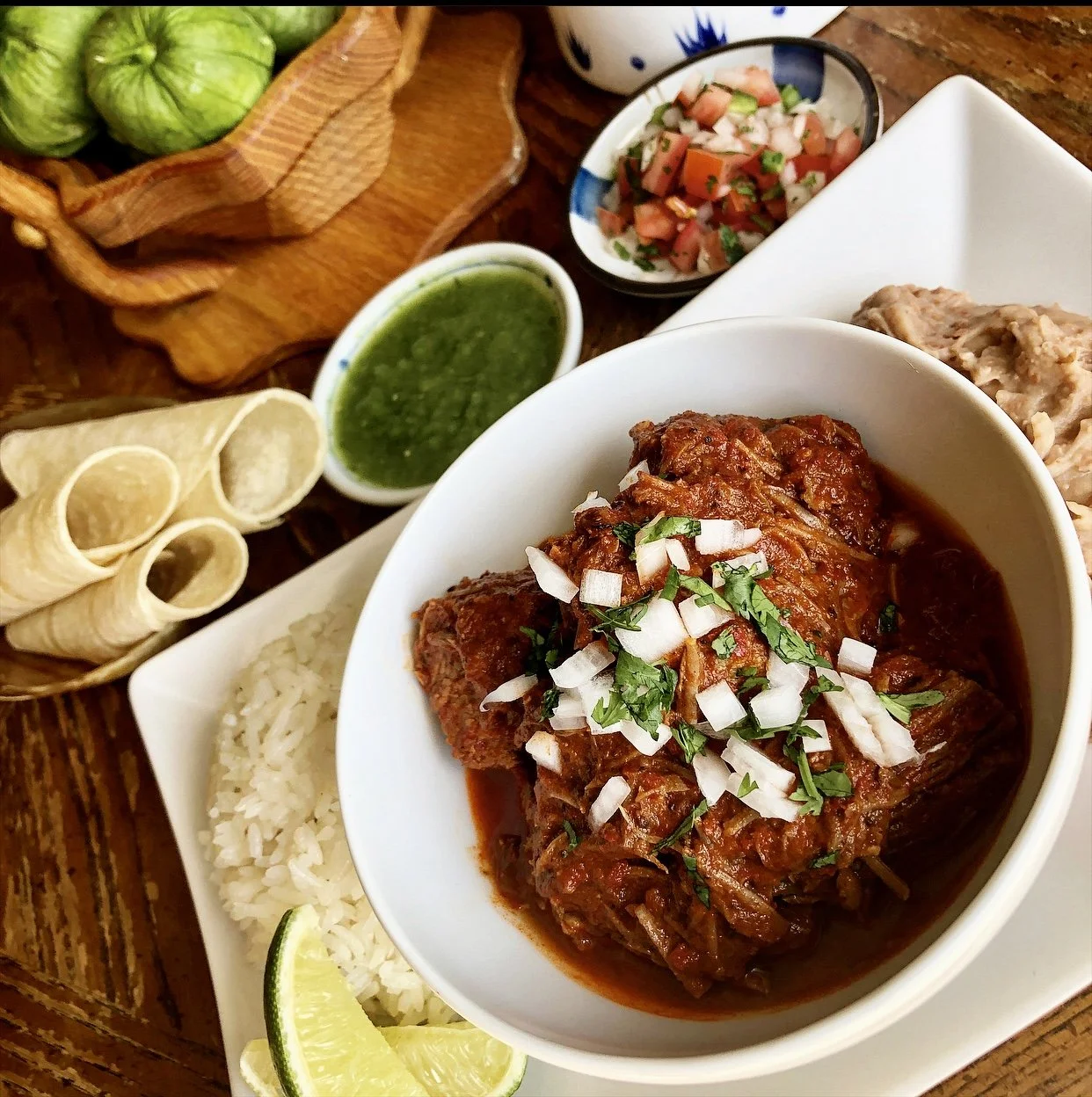 A plate of Mexican dish with shredded meat in red sauce, topped with chopped onions and cilantro, served with white rice, lime wedge, and sides of diced tomatoes, green salsa, and rolled tortillas on a wooden table.