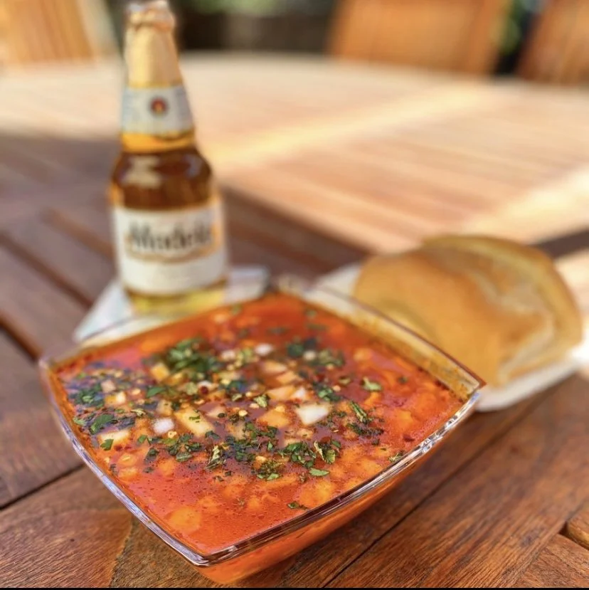 Bowl of spicy ceviche garnished with herbs on a wooden table, accompanied by bread, a bottle of Modelo beer, a roll of bread, and a napkin.
