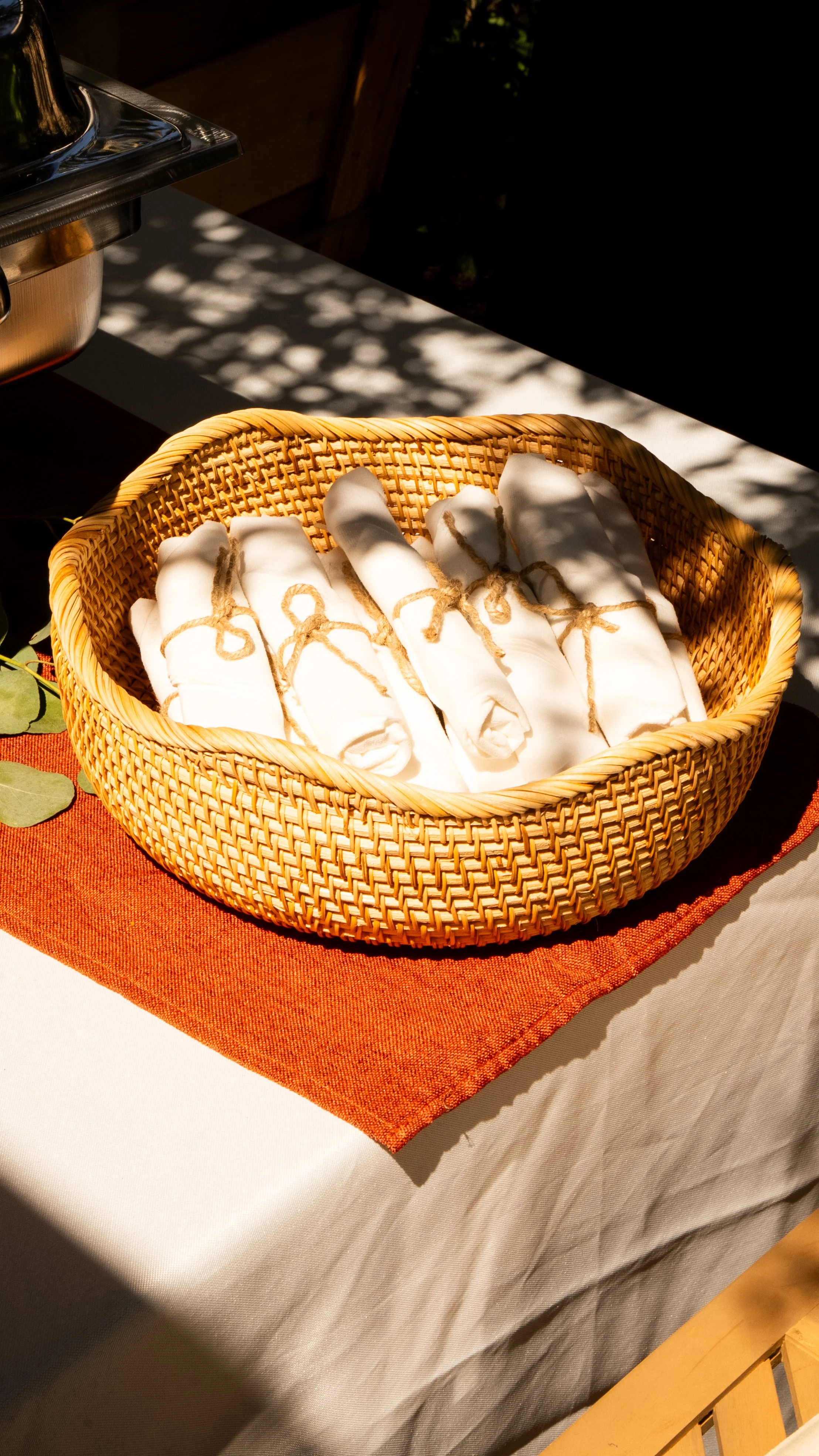 A wicker basket containing rolled white cloth napkins tied with twine, placed on a table with orange and white tablecloths, partly shaded by leaves.