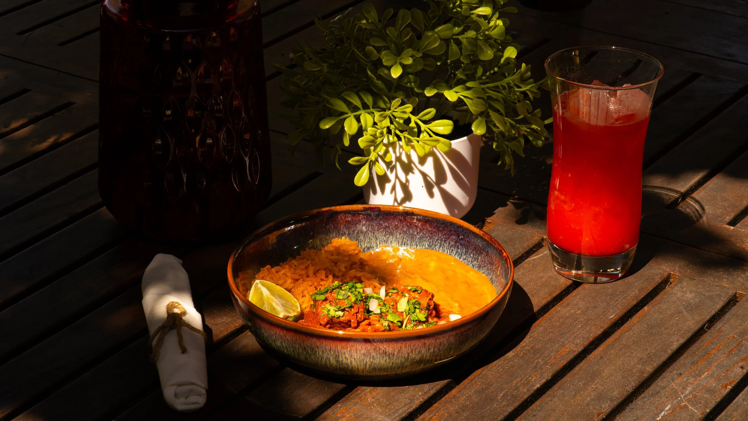 A table with a bowl of Mexican rice, a bowl of refried beans, a lemon wedge, and chopped cilantro; a tall glass of red drink, a decorative vase, and a potted green plant, all on a wooden table in sunlight.