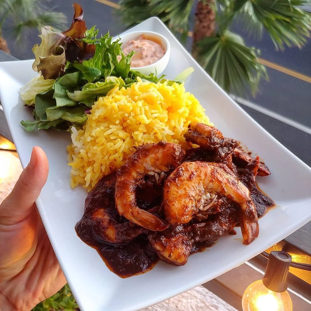 Plate with shrimp in tomato sauce, yellow rice, mixed greens salad, and a small cup of dipping sauce, outdoors near palm trees with string lights.