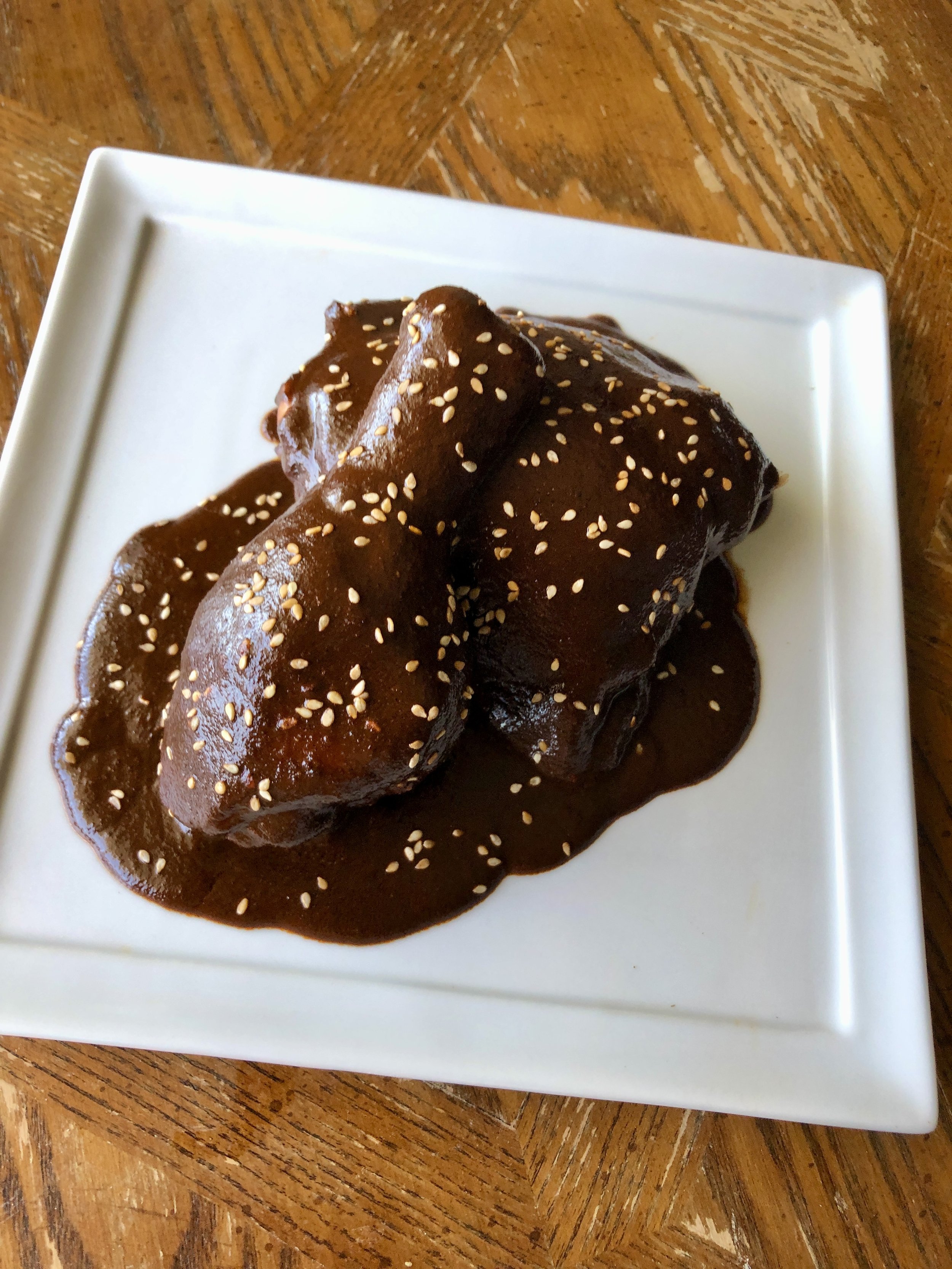 A white square plate holds a serving of chicken covered in a dark brown sauce, topped with sesame seeds, and placed on a wooden table.