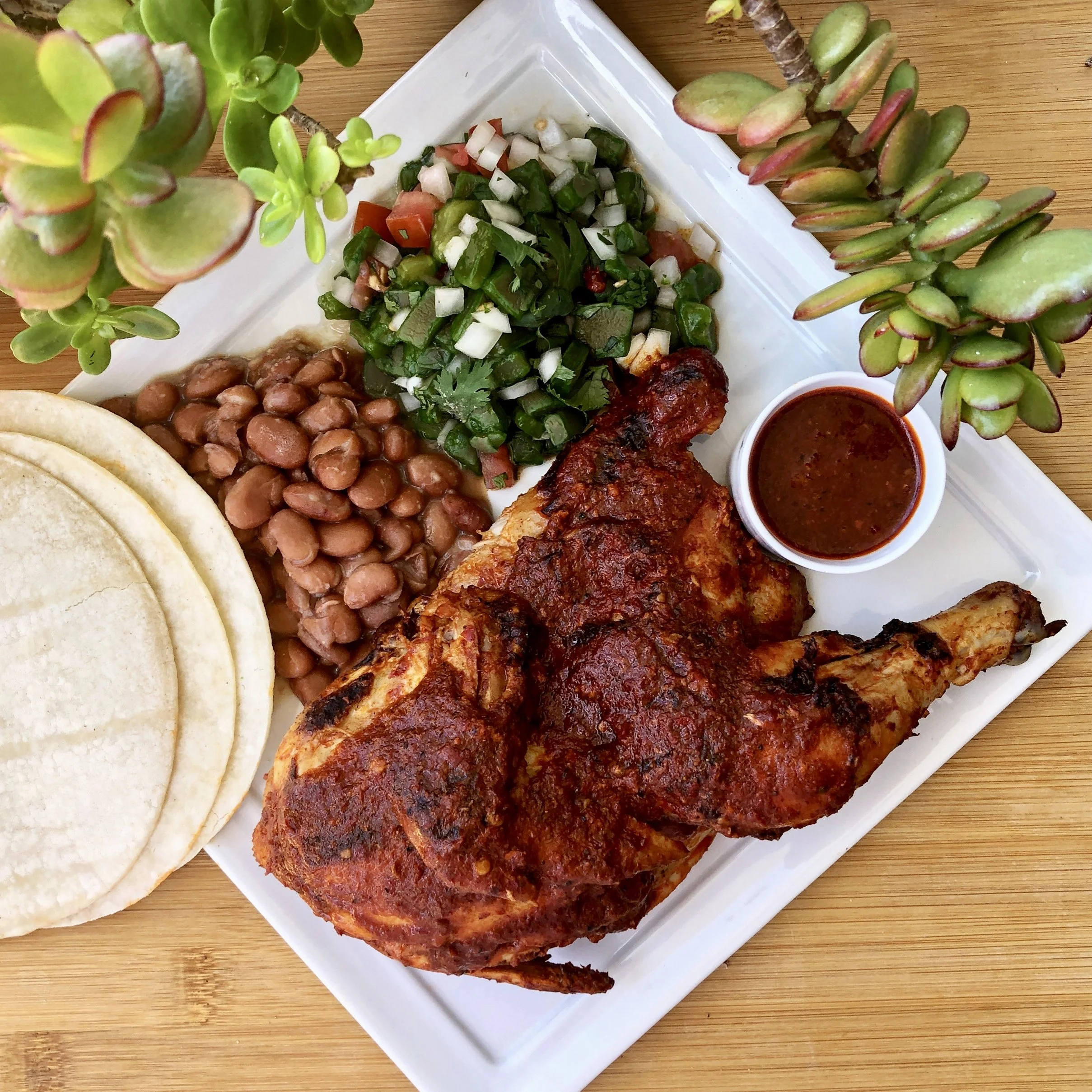 Close-up of a white rectangular plate with traditional Mexican dishes including beans, grilled meat, and vegetables, arranged on a wooden table, with green succulent plants nearby.