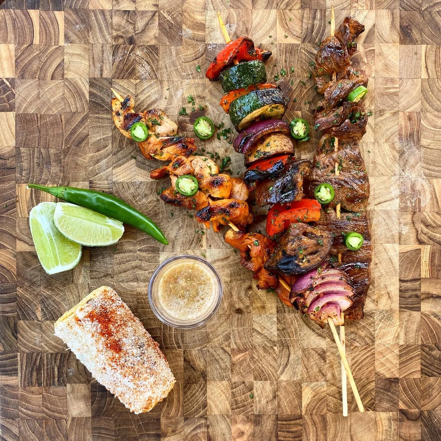 Skewers of grilled meat and vegetables, lime wedges, sliced jalapeño, a piece of crusted bread, and a small container of dipping sauce on a wooden cutting board.