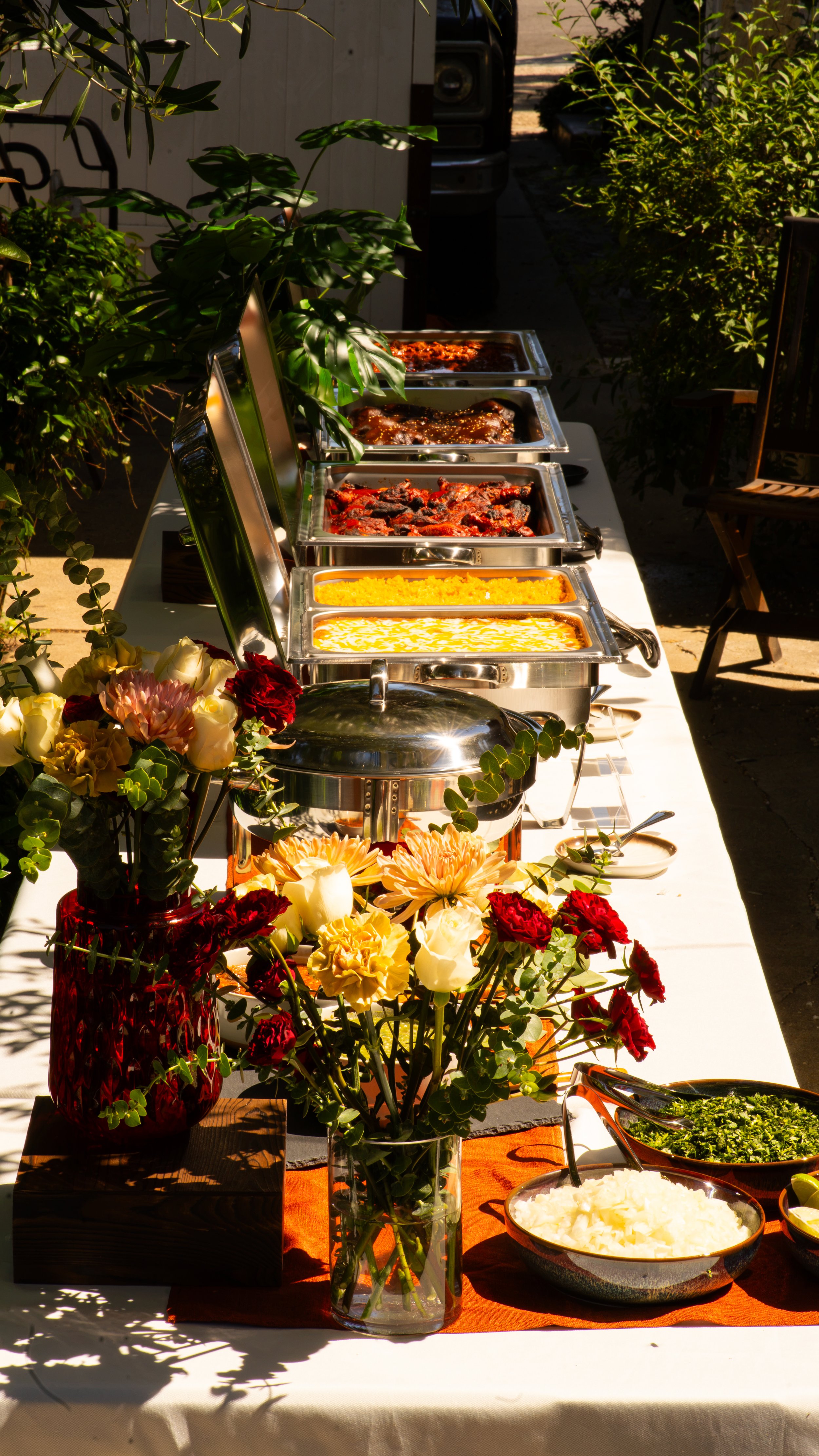 Buffet table with chafing dishes containing hot food, surrounded by flowers and greenery, set outdoors.