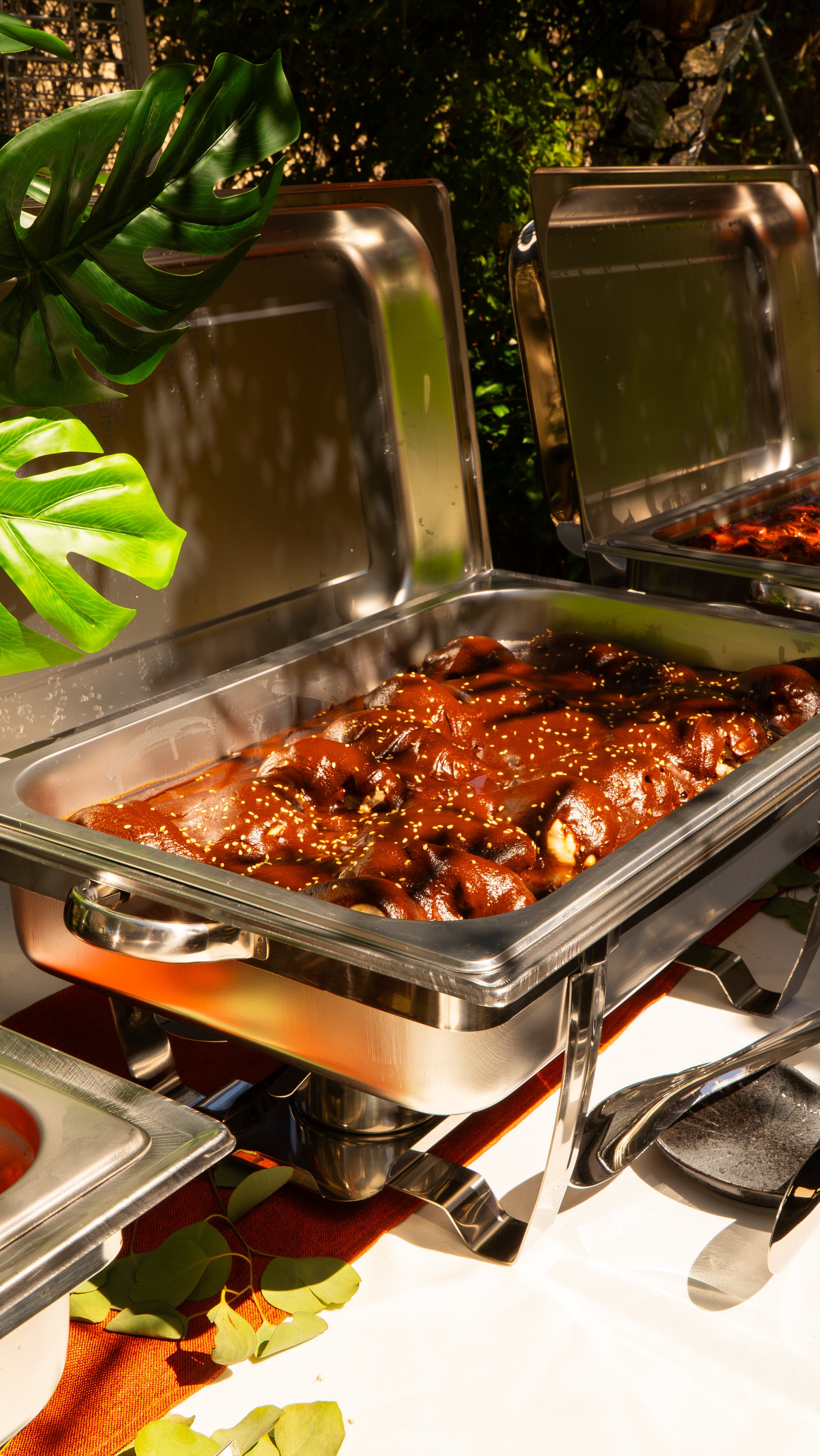 Buffet style serving tray with glazed meat or chicken, garnished with sesame seeds, set on a table with green leaves and outdoor background.