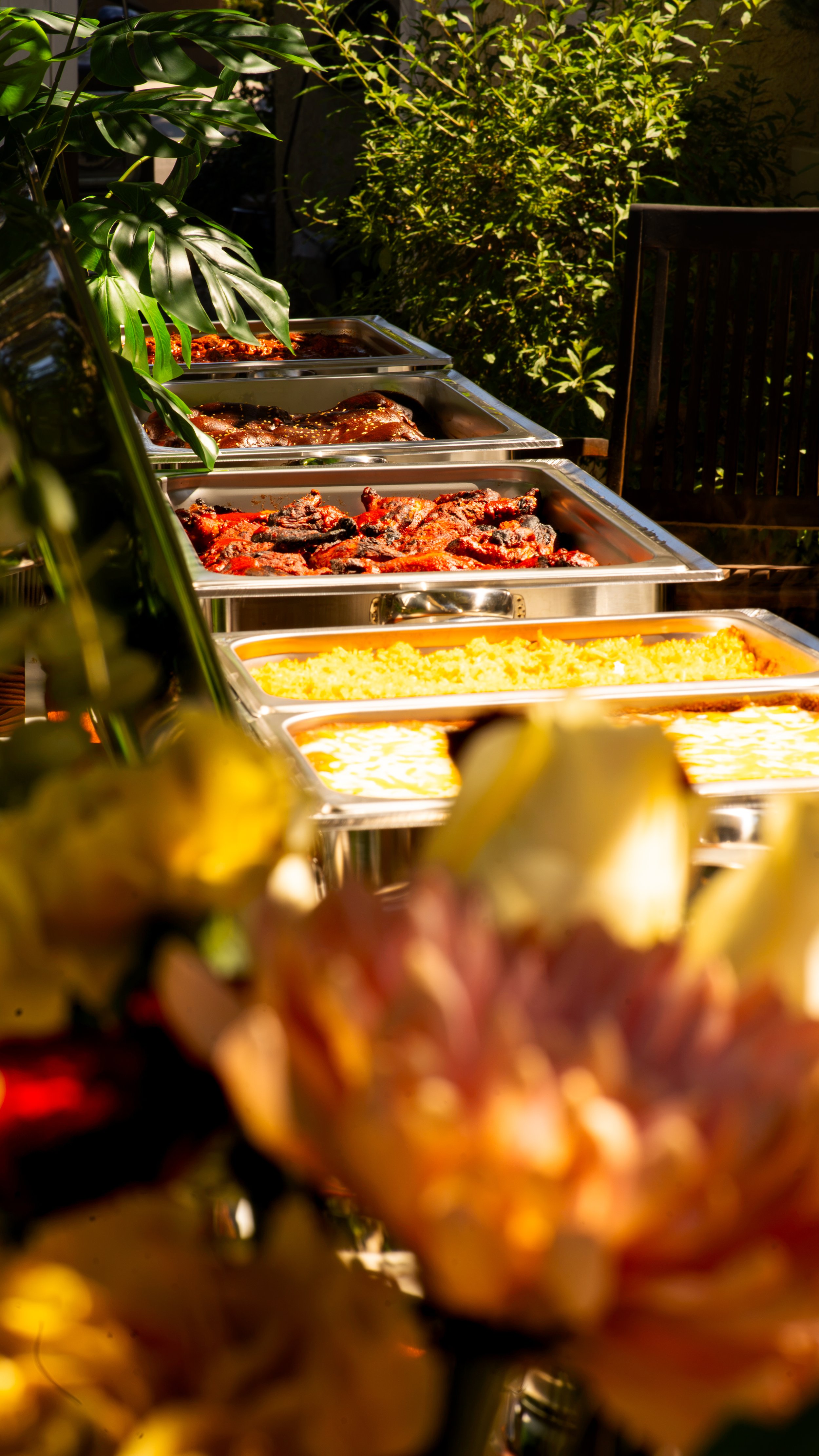 Buffet table with trays of food outdoors, partially covered by green plants, with sunlight casting a warm glow.