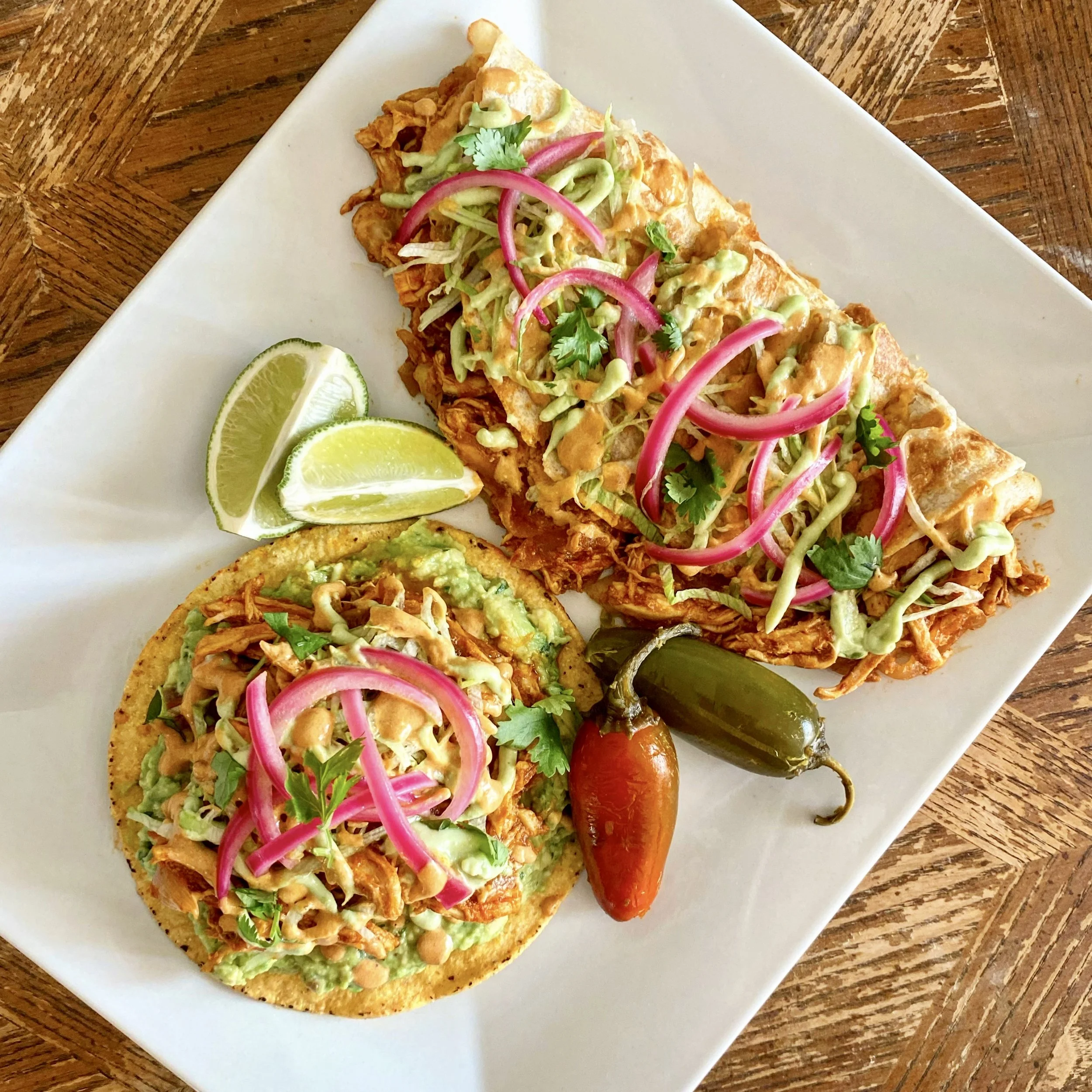 Close-up of fish tacos with shredded lettuce, pickled red onions, cilantro, drizzled sauce, and lime wedges on a white square plate on a wooden table.