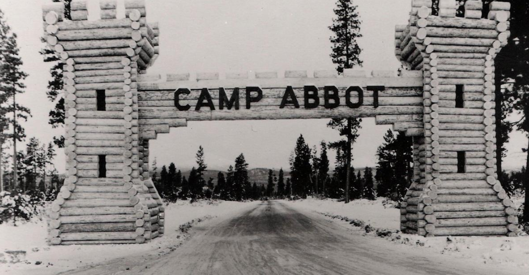 Black and white photograph of the entrance to Camp Abbot, featuring two log towers connected by a log arch with the words "CAMP ABBOT" in the center. The road leading into the camp is unpaved, surrounded by snow and trees.
