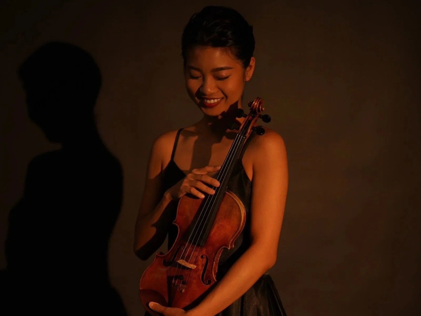 A woman with short dark hair, wearing a black dress, holding a violin, standing against a dark background, with a soft light illuminating her face and the violin, creating a shadow on the wall.