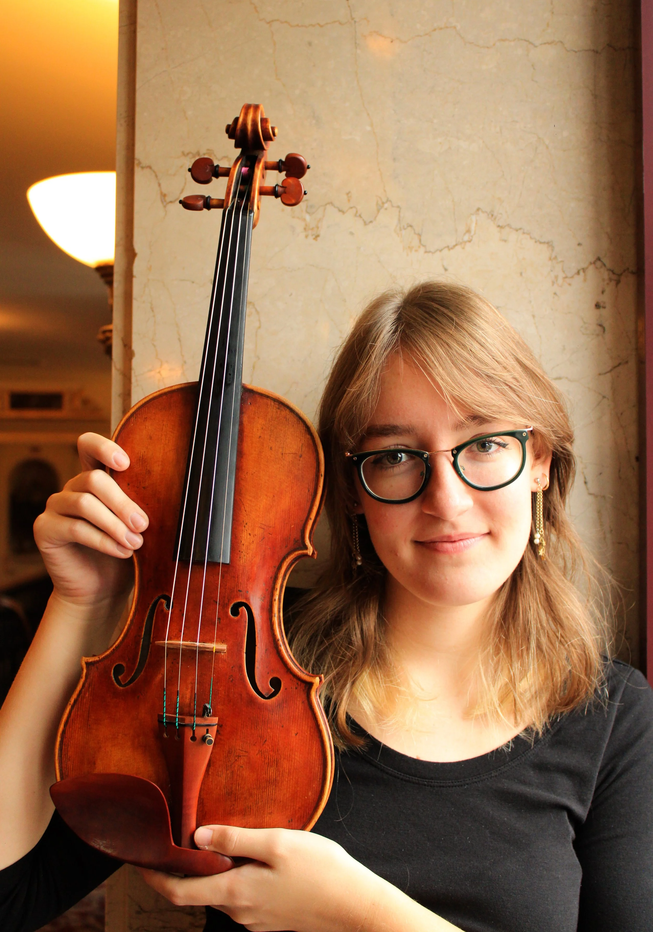 A young woman with glasses and long, light brown hair holding a violin in front of a textured beige wall.