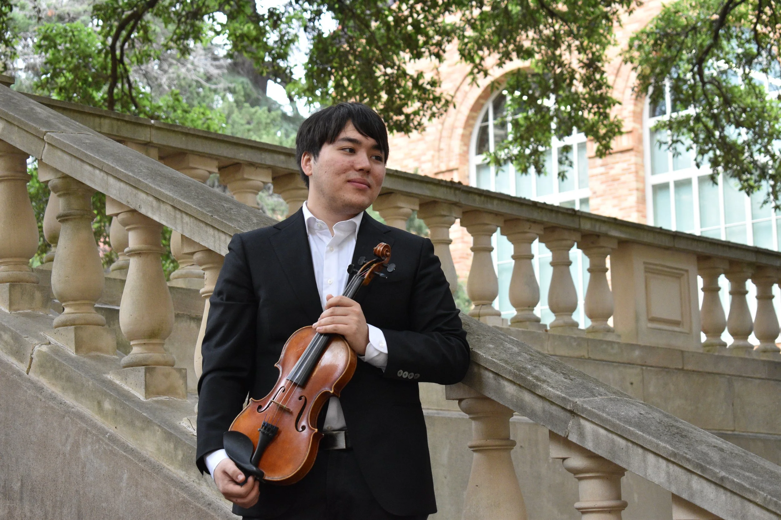 A young man in a black suit with a white shirt holding a violin, standing on outdoor stone stairs with a brick building and green trees in the background.