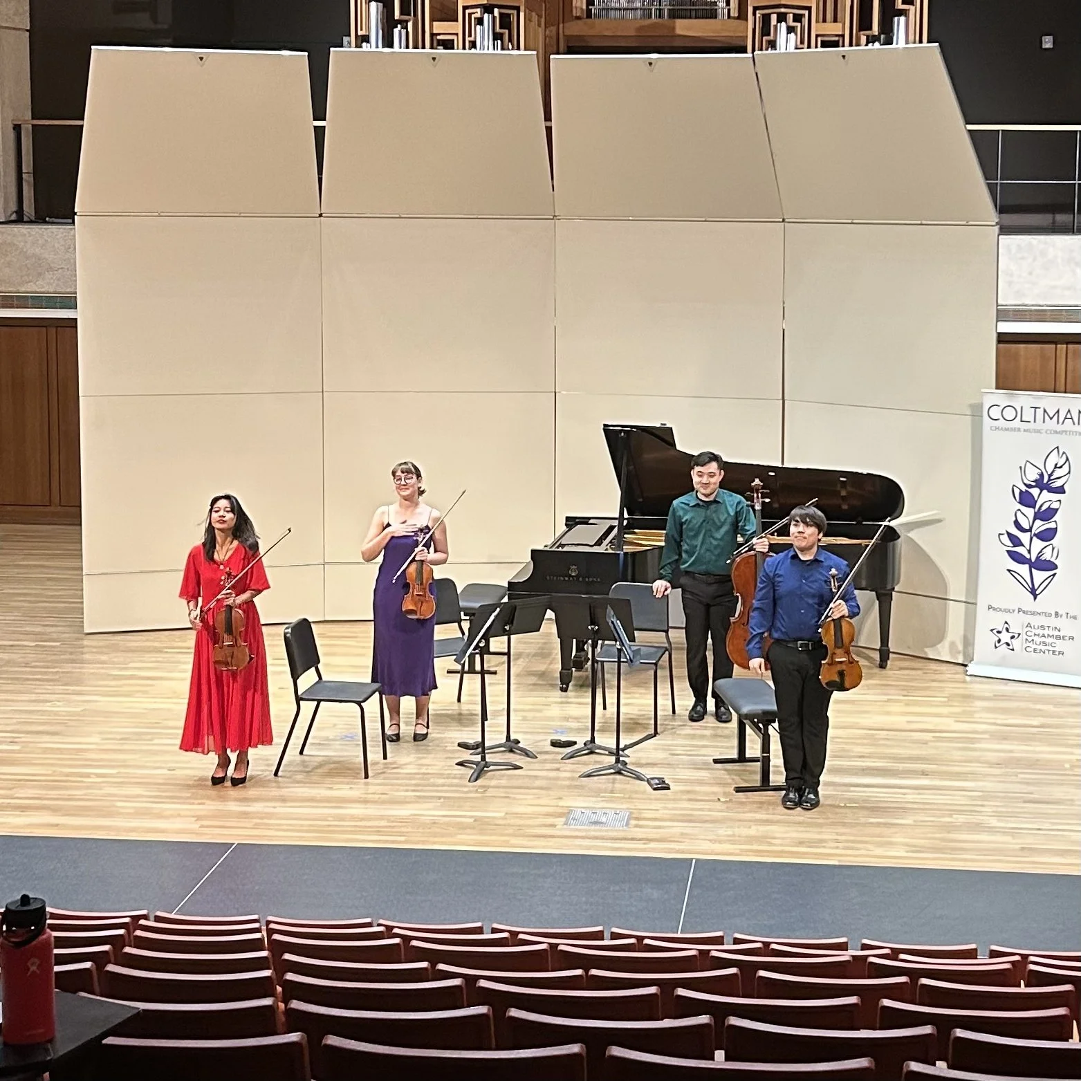 Four musicians holding their instruments on stage after a concert for the Coltman Competition, with a grand piano behind them and a sign for the Austin Chamber Music Center.