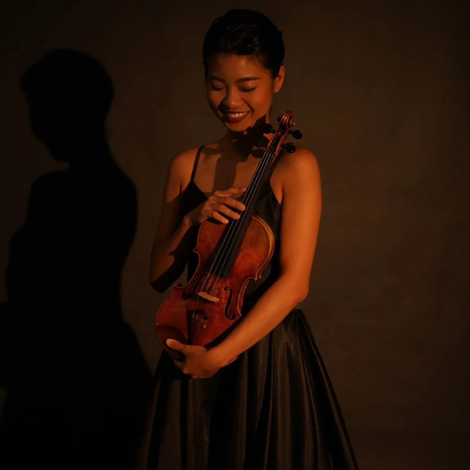 A woman with short hair in a black dress holding a violin and smiling against a dark background with a shadow cast on the wall.